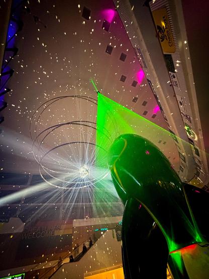 Blick nach oben. Eine große, grüne Rakete, die Fairy Dust, steht in der bunt beleuchteten Eingangshalle des Hamburger CCH. Ein grüner Laserstrahl leuchtet von der 25 Meter hohen Decke, der durch die vernebelte Luft schneidet, und einen Beam-Effekt um die Rakete herum erzeugt. Der verspiegelte Discoknoten unter der Decke reflektiert weiße Strahlen, die tanzende weiße Punkte wie Sterne an die Decke werfen. Er wirkt so wie ein Zentralstern in der Chaos-Galaxie. Im Hintergrund erkennt man vage das bunte Banner „All Creatures welcome“. Nicht erkennbare Kreaturen fahren in der Ferne eine Rolltreppe hinunter.