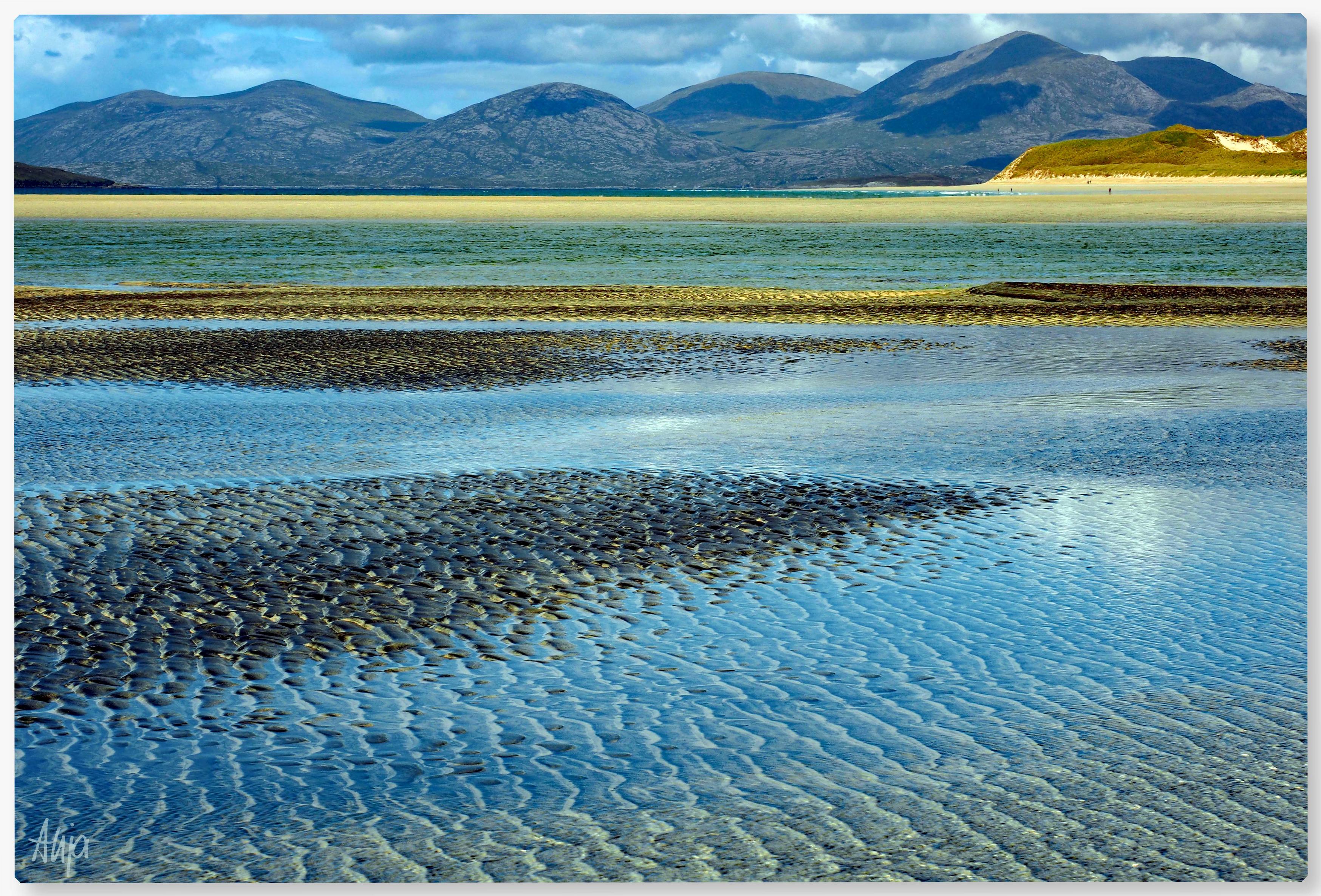 Ebbe in der Bucht bei Seilebost, Harris, Schottland: Das verbliebene Wasser in den Rippeln leuchtet in den unterschiedlichsten Blau-und Grüntönen, die höheren Stellen sind dunkler, fast blau-schwarz. Im Hintergrund ein Sandstreifen vor Dünen, auf die das Sonnenlicht fällt. Winzige Silhouetten, Menschen, die dort spazierengehen. Und die Hügel von North Harris, über denen Regenwolken hängen.
Low tide in the bay at Seilebost, Harris, Scotland: the remaining water in the ripples glows in various shades of blue and green, while the higher areas are darker, almost blue-black. In the background, a strip of sand in front of dunes bathed in sunlight. Tiny silhouettes, people walking there. And the hills of North Harris, with rain clouds hanging over them.