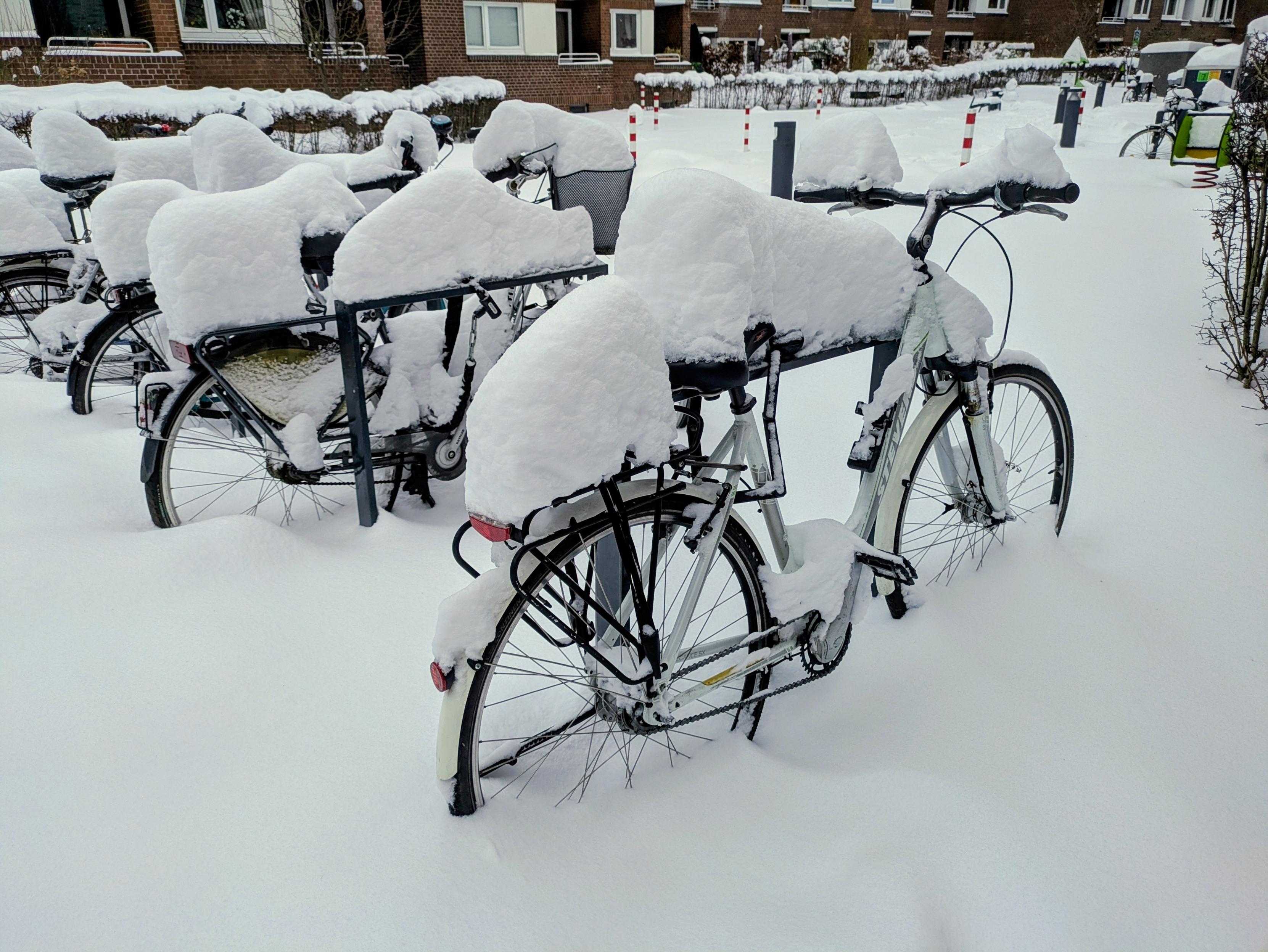 Mehrere Fahrräder vor einem Wohnblock an mehreren Fahrradbügeln, schneebedeckt, inmitten von frischem Schnee.