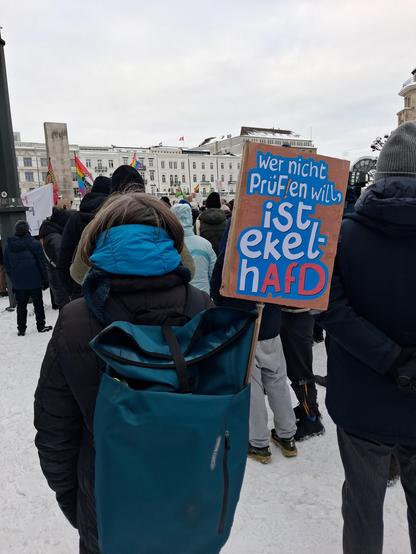 Mensch (weiblich gelesen) von hinten, dick eingepackt. Aus mehreren Jacken und einem Strickschal gucken oben nur ein paar Haare raus. Die Person trägt einen großen Rucksack, aus dem ein Schild an einer Bambusstange ragt: Wer nicht PrüF!en will, ist ekelhAfD. (Der Hintergrund der Schrift ist in cyanblau, die letzten 3 Buchstaben hellrot, eine Anspielung an die Rechtsaußenpartei). Um die Person herum im Schnee stehen andere Menschen, es liegt Schnee, man sieht noch andere Flaggen (Regenbogen) und…