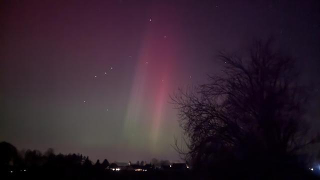 Nordlicht. Der Himmel direkt über dem Horizont schwach grün, in der Mitte des Bildes ein breiter roter Streifen vertikal nach oben weg. Rechts im Bild die Silhouette eines alten Baumes. Unterhalb des Horizonts ein paar Lichter des Dorfes.