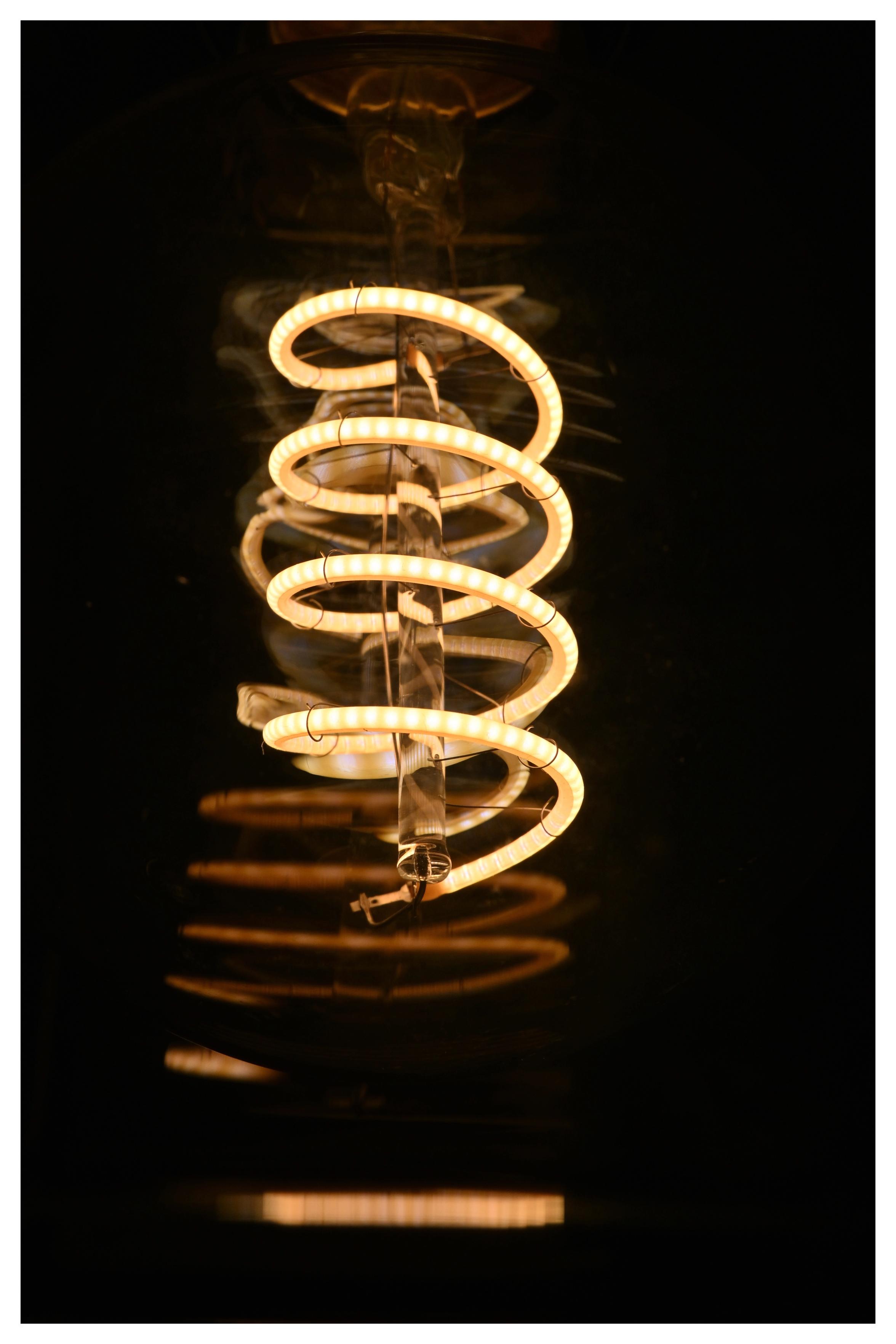 Ein LED-Leuchtmittel in Form einer Glühlampe mit einer in Gelb leuchtenden LED-Spirale, das Glas der Leuchte erzeugt sanfte Reflexionen in Orange. Drumherum dunkler Hintergrund.