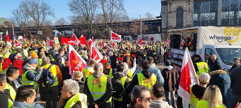 Versammlung vieler Menschen unter freiem Himmel. Viele haben Fahnen der Gewerkschaft Verdi und oder tragen gelbe Westen mit Verdi-Aufdruck. Das Tageslicht ist hell und freundlich, im Hintergrund ist eine Schienenbrücke, unbelaubte Bäume und rechts der Bahnhof Dammtor zu erahnen. Zwischen Menschenmeer und Bahnhof steht rechts ein Lautsprecher-LkW. Die Plane ist geöffnet bei der der Menge zugewandten Seite, eine Person ist auf dem LKW zu erahnen.
