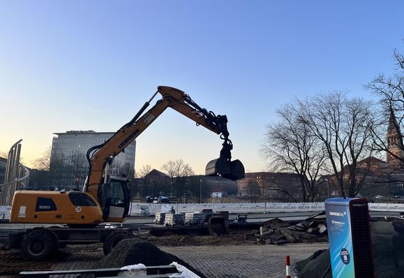 Eine Baustelle auf einem Platz in morgendlich tief stehender Sonne. Im Hintergrund ein Park mit See, dahinter das Kieler Rathaus und weitere prunkvolle Häuser.