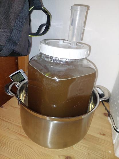 Blick auf ein 4-eckiges Gärgefäß, stehend in einem Topf auf einen Holzschrank, im Hintergrund ein Steuergerät und ein Rucksack, hängend an dem Regal links. Rechts sind Kabel zu sehen und ein wenig von der Gärbox aus Styropor. Das Gärgefäß ist mit einem weißen Deckel verschraubt, in dem ein dreiteiliges Gärröhrchen steckt. Im Gärgefäß befindet sich eine braune Flüssigkeit.