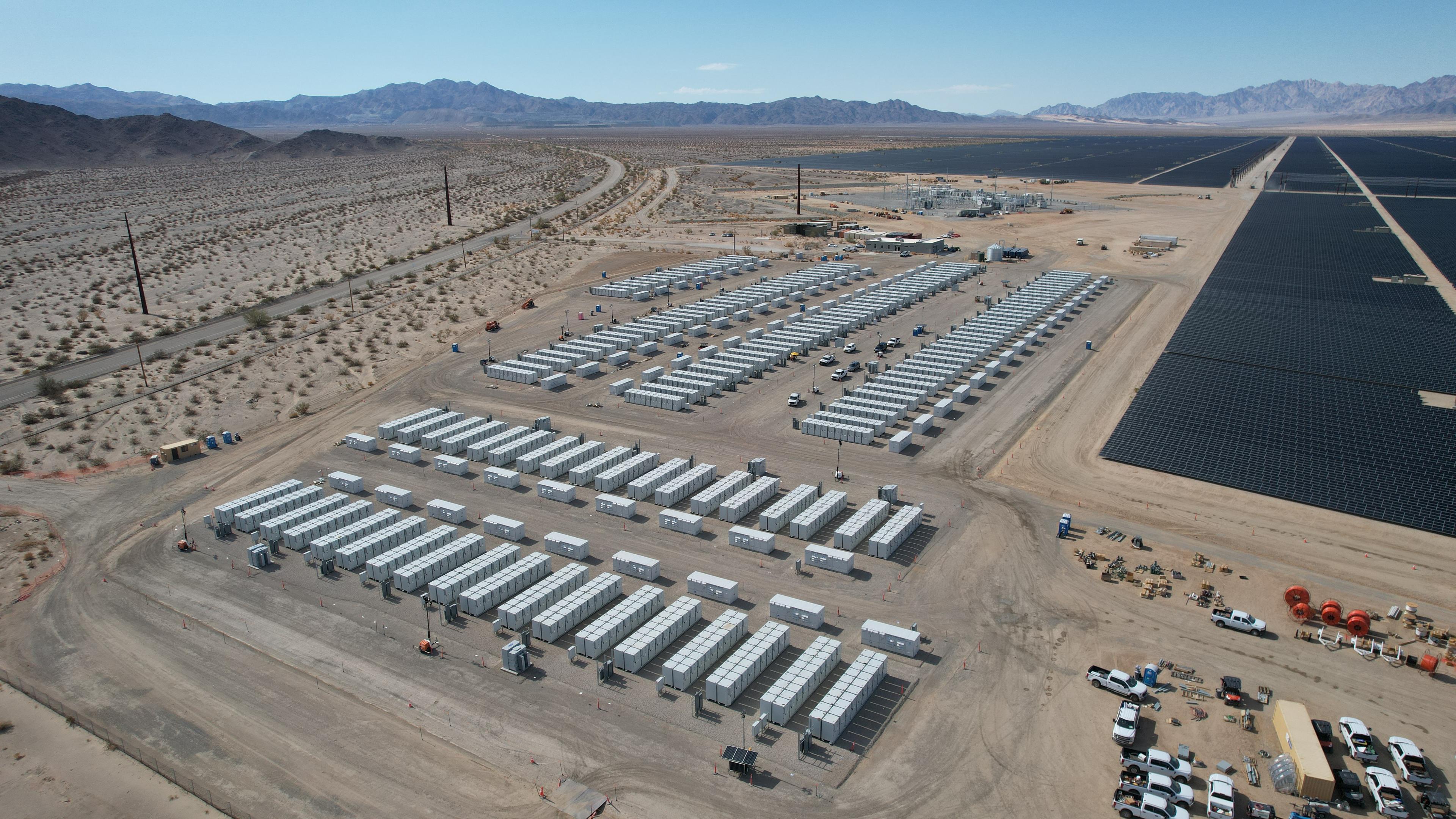 A vast array of shipping containers for battery storage is visible in a desert landscape, adjacent to solar panels in the distance.