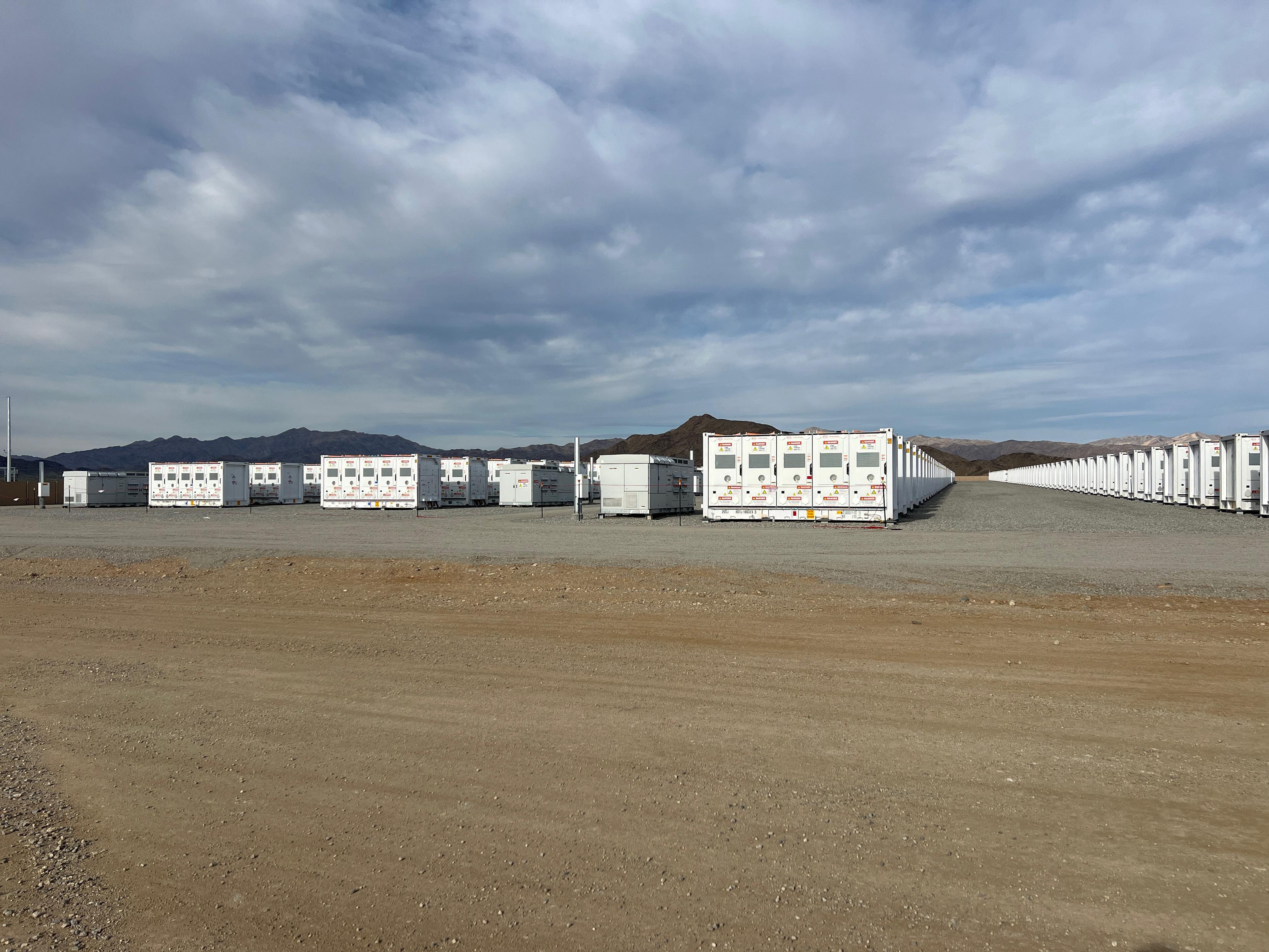 Rows of large white battery storage units stretch across a gravel site, with mountains and a cloudy sky in the background.