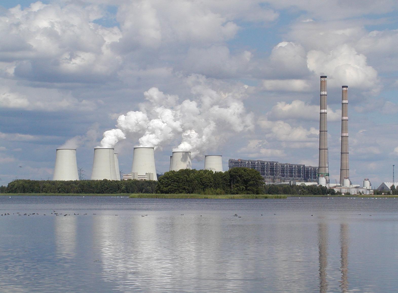 Cooling towers emit steam near a lake, with industrial buildings and smokestacks visible against a cloudy sky, highlighting energy production.