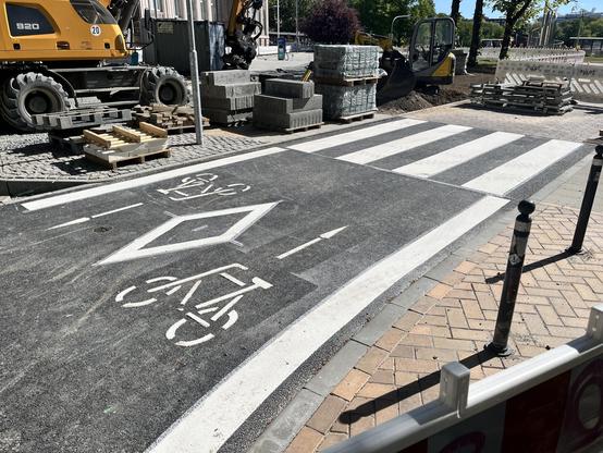 Foto einer Baustelle. Auf einer breiten Asphaltbahn sind ein querender Zebrastreifen und Fahrradsymbole in beide Richtungen der Bahn angebracht. Eine Markierung kündigt vor dem Zebrastreifen die Installation eines Pollers an.