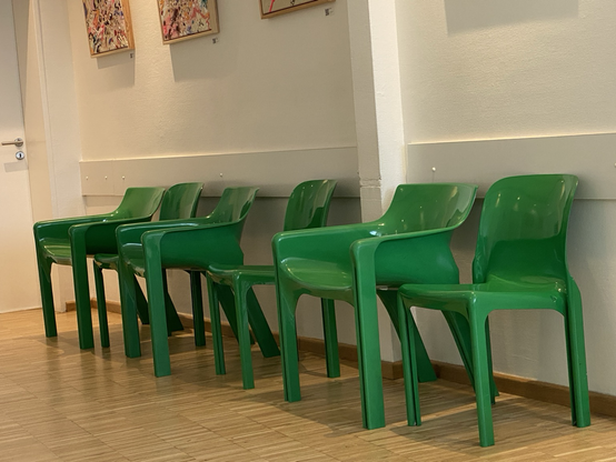 6 knallgrüne Monoblock-Design-Kunststoffstühle. Einige mit Armlehnen, einige ohne. Auf Parkett vor weißer wand mit Bildern darüber (abstraktes Geklekse, schön)