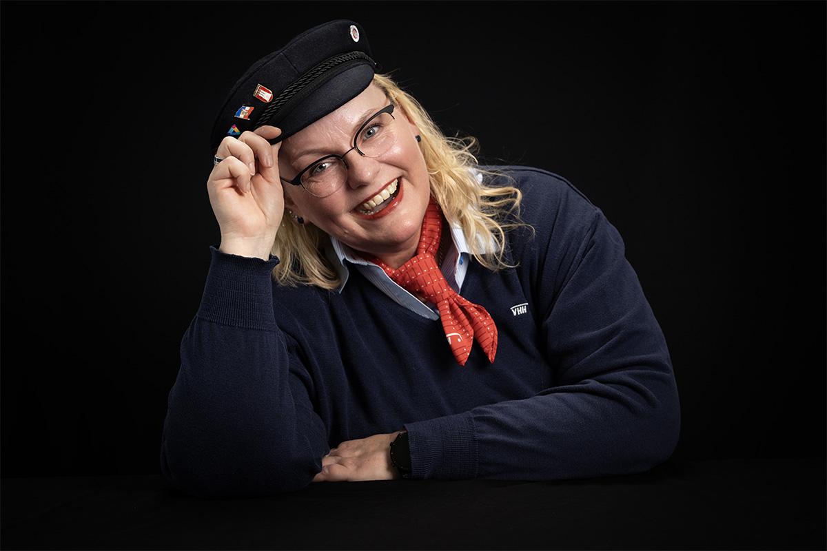 Frau Müller MeierSchulze vor einem schwarzen Hintergrund. Ich trage Hemd,und dunkelblauen Pullover, dazu ein rotes Halstuch. Ich lehne mich nach links (vom Bildbetrachter aus gesehen) habe den rechten Arm aufgestellt und lüfte leicht meinen dunkelblauen Elbsegler auf dem Kopf . 
Ich lache mit geöffneten Mund direkt in die Kamera.
