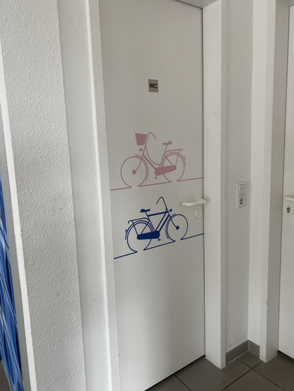 auf einer weißen Tür ist oben ein rosa Damen-Fahrrad abgebildet, also ohne Stange, und unten ein blaues Herren-Fahrrad, also mit Stange.