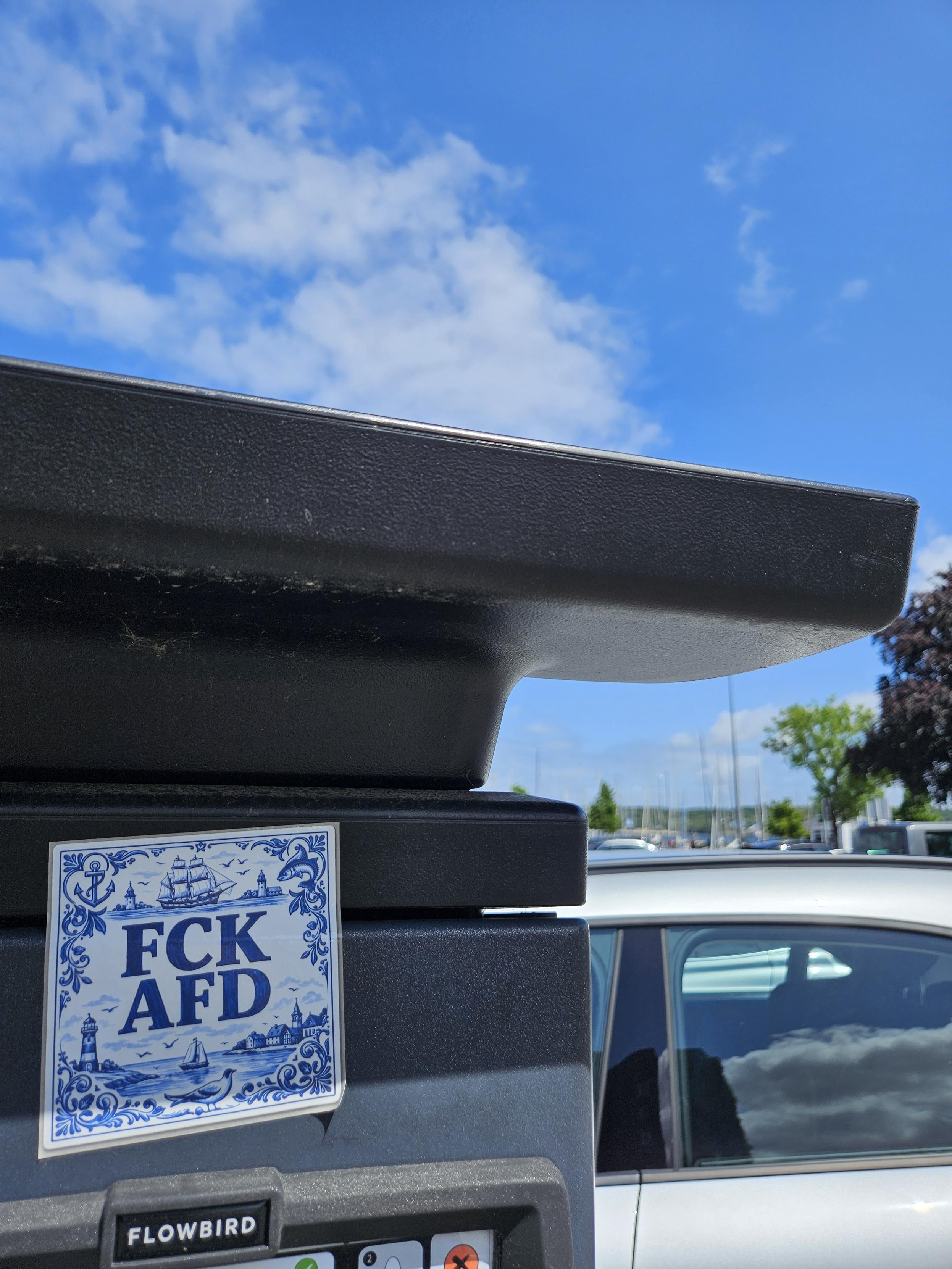 Ein maritim gestalteter FCK AFD Aufkleber an einer Parkticketsäule in Kiel, im Hintergrund sind Segelmasten zu erkennen.