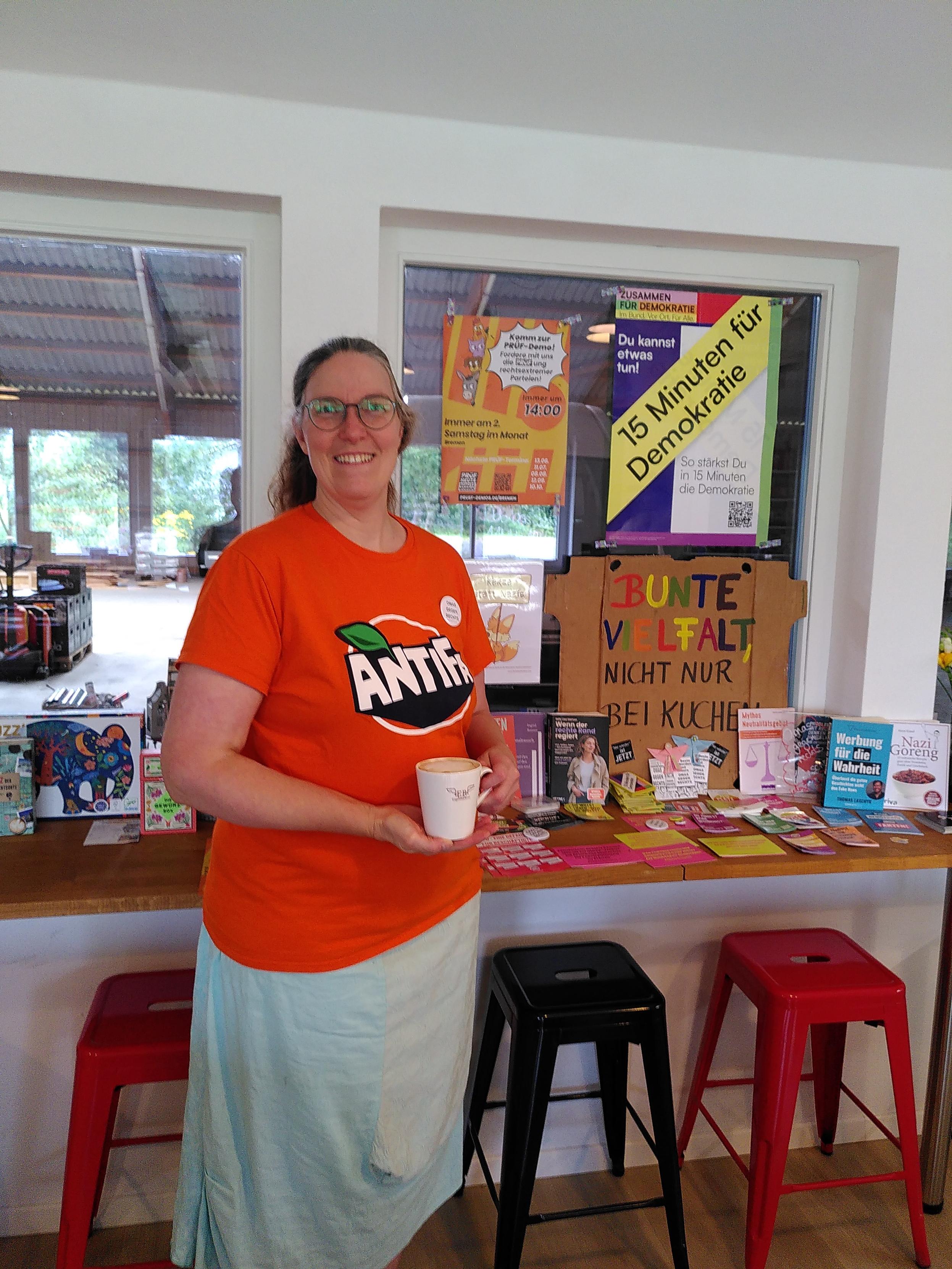 Ich stehe vor meinem Demokratiefenster im Café. Ich trage einen hellgrün-türkisen Rock und ein quietsch orangefarbenes T-Shirt mit dem des Fanta-Logos nachempfundenen Aufdrucks ANTIFA. Ich halte einen weißen Kaffeebecher mit dem Logo meines Cafés und lächele in die Kamera. 
Im Demokratiefenster hängen verschiedene Plakate rund ums Thema Demokratie und Prüf-Demos, davor liegen diverse Postkarten, Handzettel und andere Informationsmaterialien.