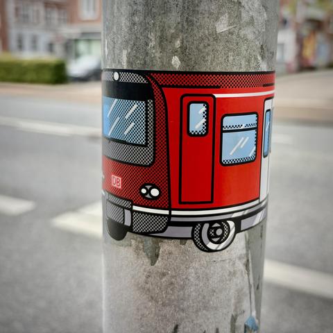 Ein Aufkleber stellt eine S-Bahn dar. Er klebt ab einem Straßenschildpfahl.