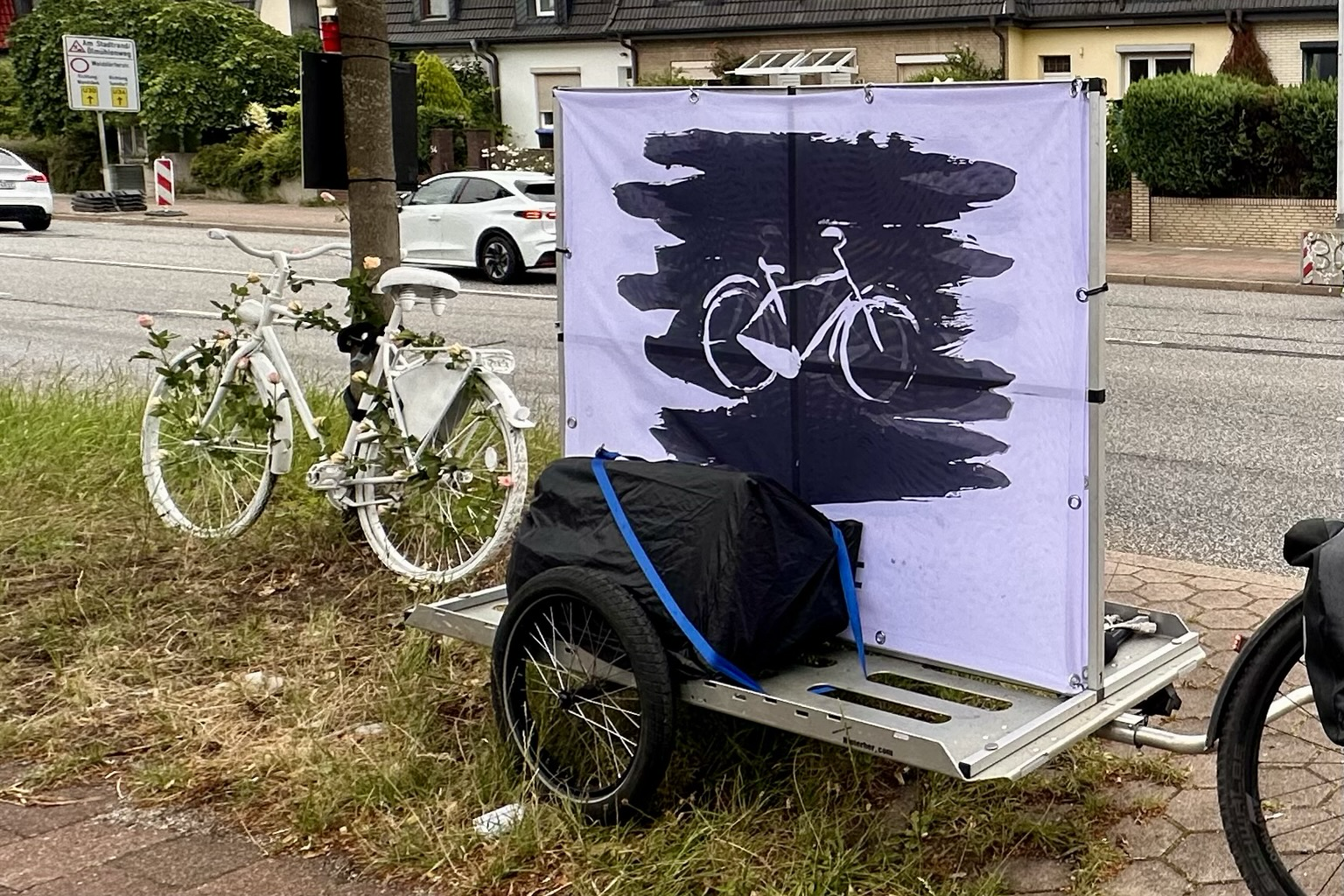 Transportanhänger mit Banner StopKillingCyclists u Lautsprecher. Ghostbike lehnt daneben am Baum.