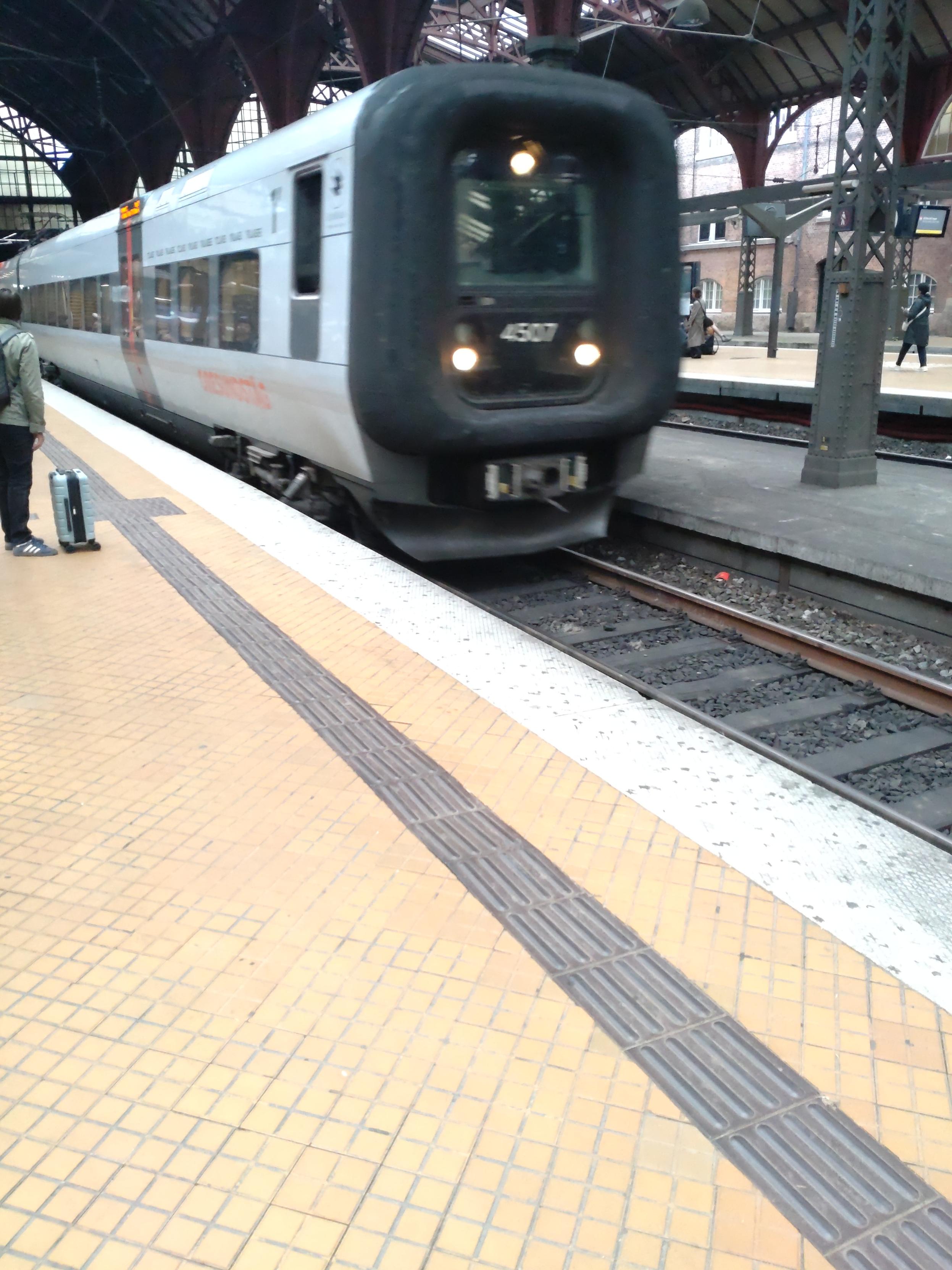 Ein Øresundtåg fährt am Bahnsteig ein.   Am Gleis wartet eine verhaltene Menge an Pendlern und Flugreisenden.