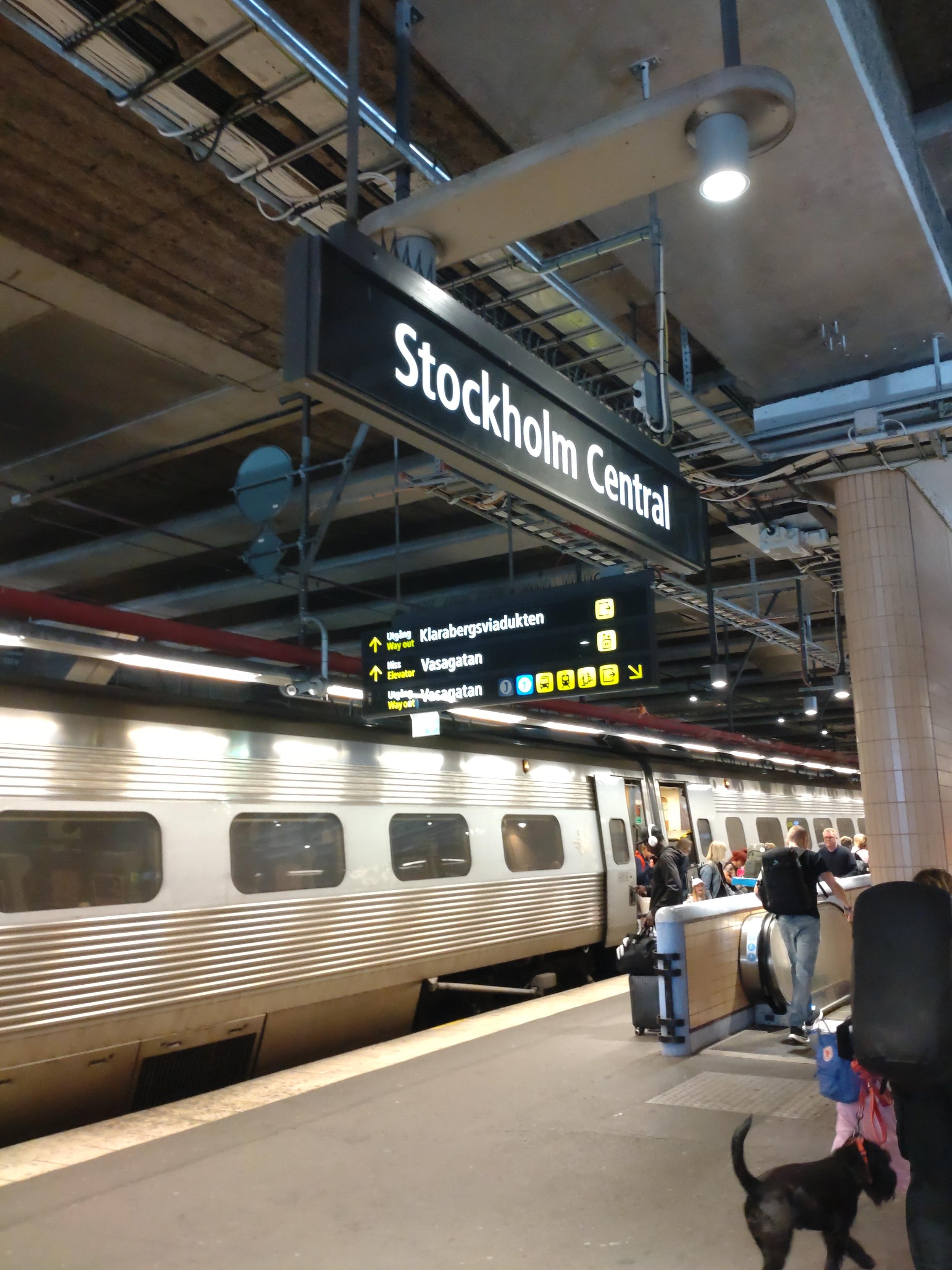 Ein Bild vom Bahnsteig. An der Decke ist ein beleuchtetes Schild Stockholm Central angebracht. Der X2000 steht im Hintergrund am Bahnsteig. Reisende gehen ihrer Wege.