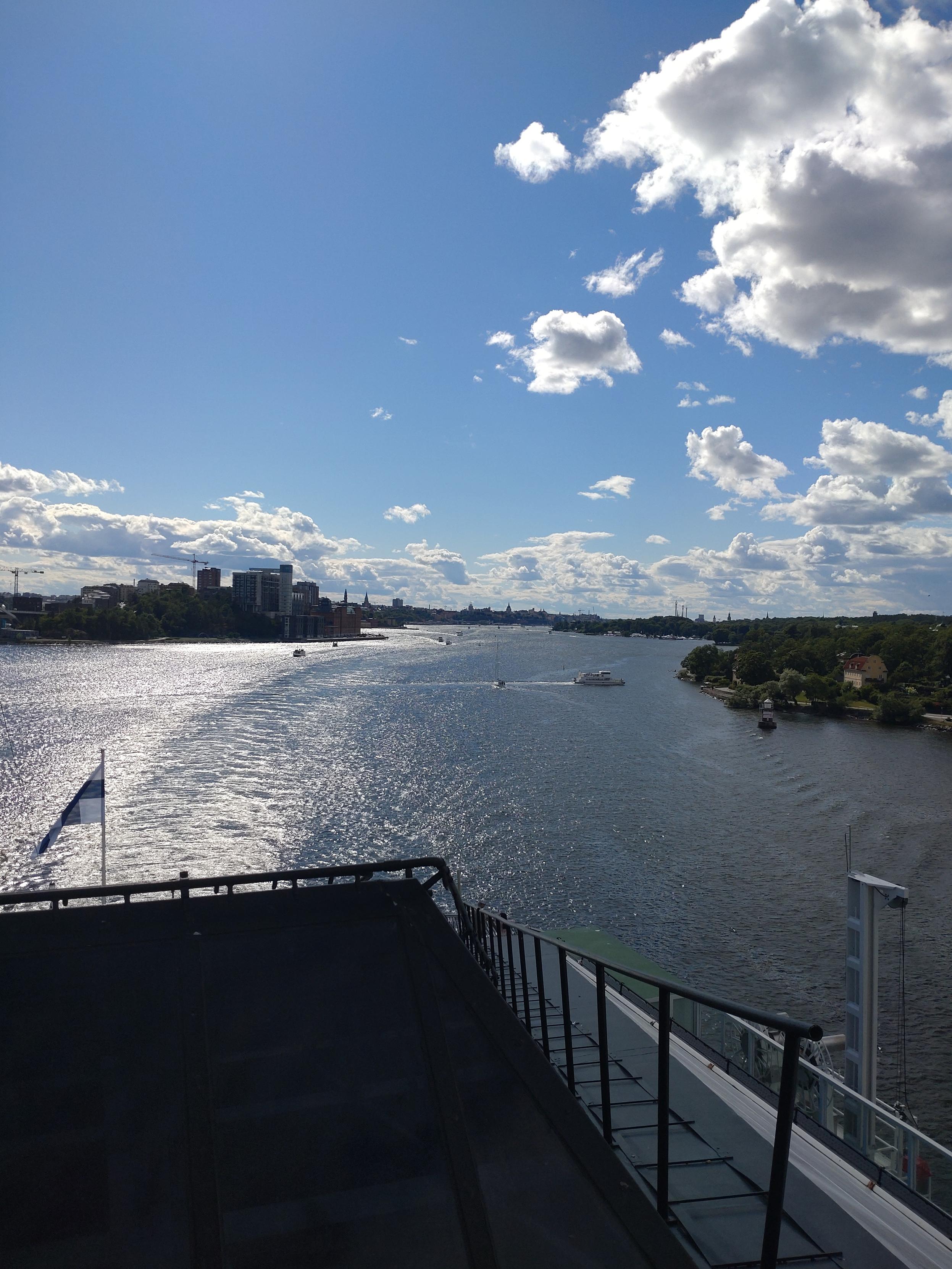Blick auf das Heck des Schiffes mit einer großen Finnischen Fahne. Im Hintergrund ist die Stadtsiluette der Stockholmer Innenstadt zu erkennen. Das Wetter hat blauen Himmel mit Schönwettet Wolken.