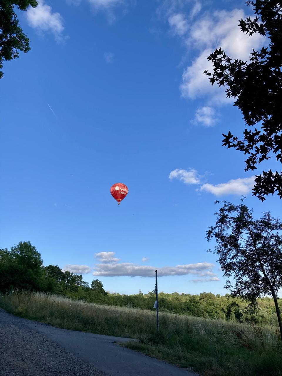 Am blauen Himmel, der nur am Rand des Bildes und in der Ferne von einzelnen Wolken bedeckt ist, schwebt ein roter Heißluftballon über einer Wiese. Das Gelände fällt nach rechts leicht ab. Hinter der Wiese stehen viele Bäume, vor der Wiese verläuft in der linken unteren Bildhälfte ein befestigter Weg. Im Vordergrund ragen von links und rechts Bäume teilweise ins Bild.