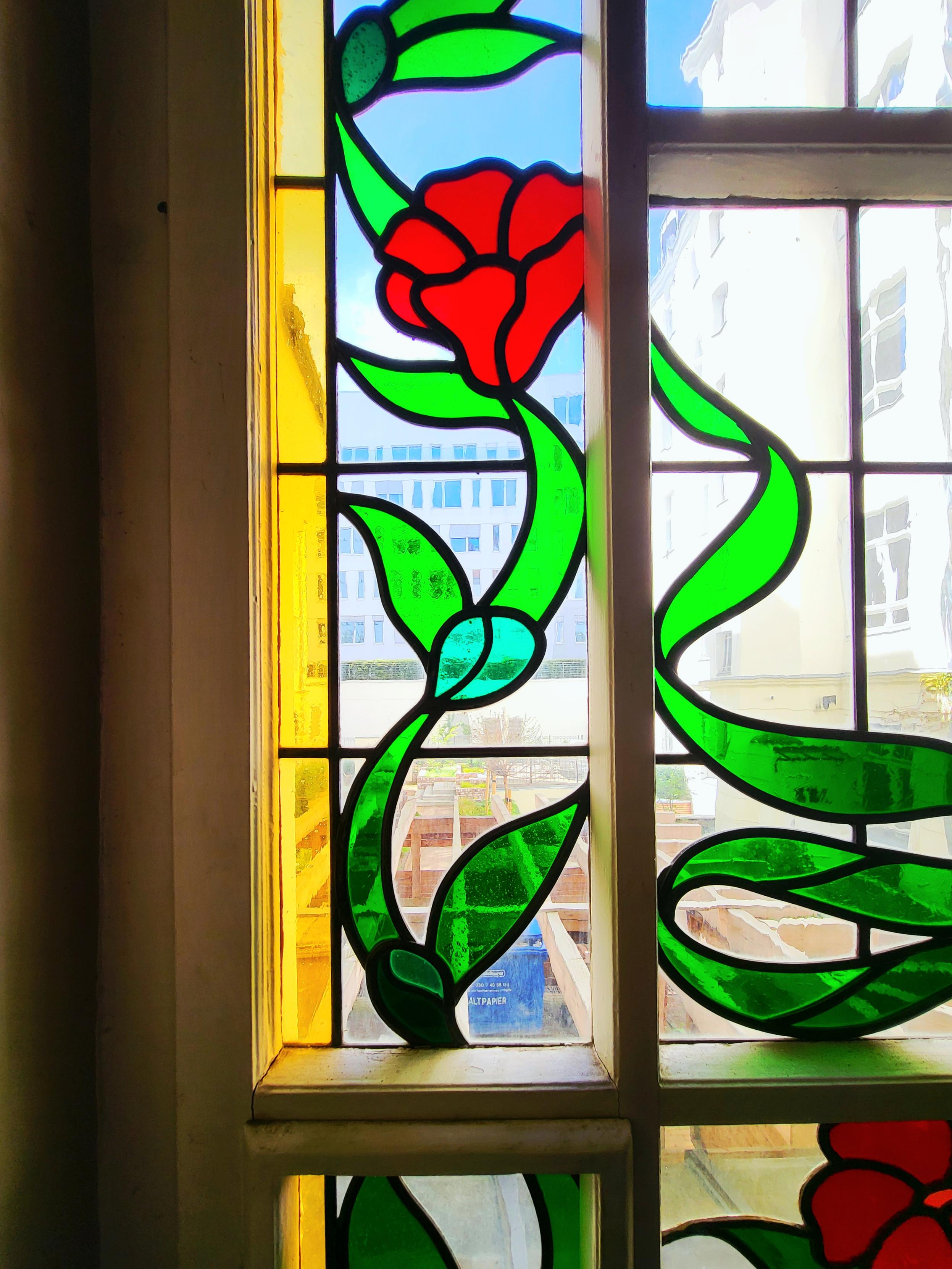 Buntes Bleiglasfenster mit grünem floralem Muster und roten Blüten im Treppenhaus eines Berliner Altbaus aus dem 19. Jh, an Jugendstil abgelehnt. Durch das Fenster fällt der nicht ganz klare, unterbrochene Blick durch die verschiedenenfarbigen Glasfacetten in grün, rot, gelb und klar-transparent auf einen weitläufigen Hinterhof mit Pergola über dem Müllplatz und auf einen Seitenflügel. Im fernen Hintergrund ein weißes Gebäude vor blauem wolkenlosen Himmel.