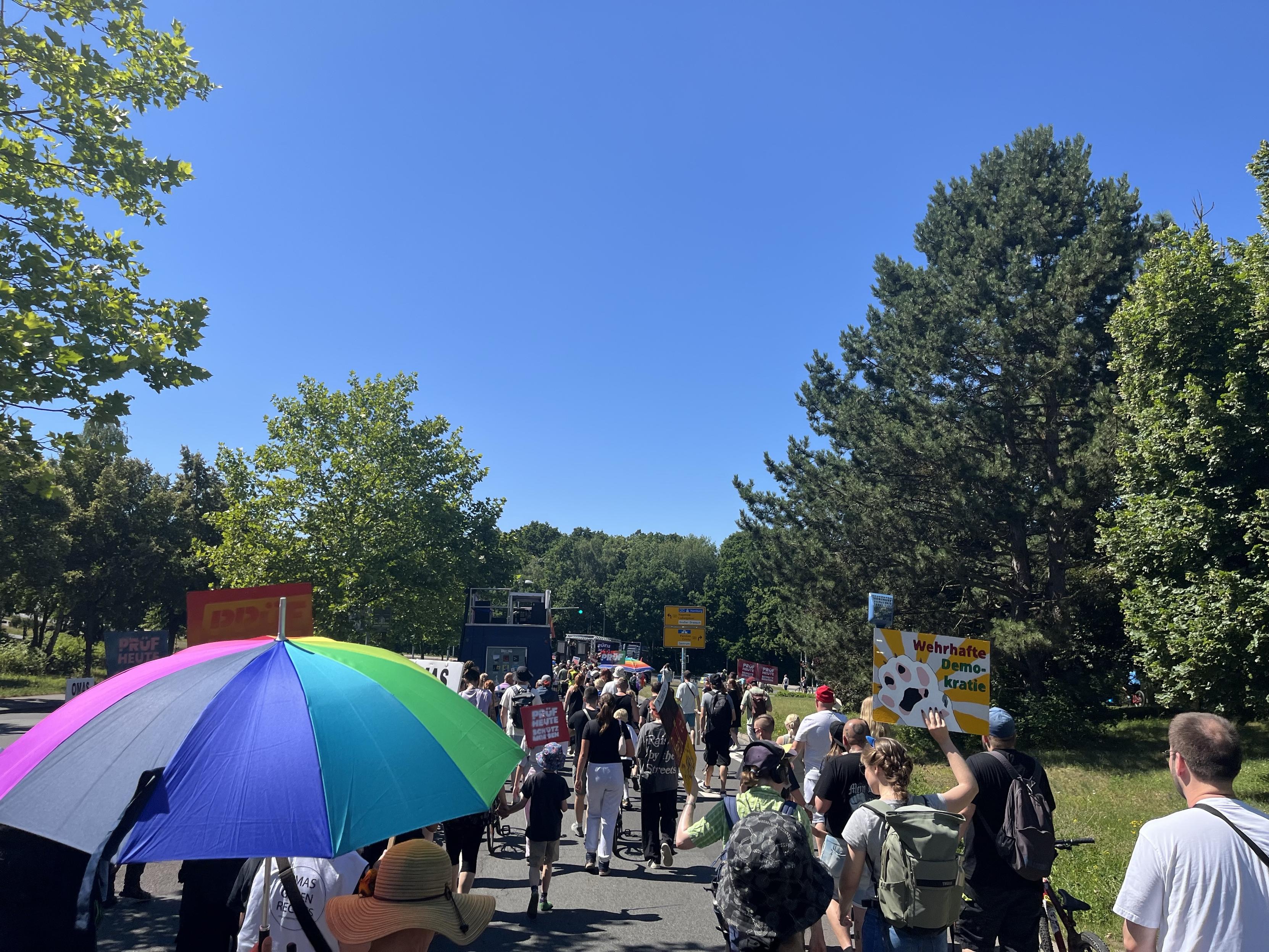 Eine vielfältige Menge nimmt an einer Demonstration unter einem klaren blauen Himmel teil. Manche Menschen tragen bunte Regenschirme und Schilder, während Bäume den Weg säumen. Die Atmosphäre ist lebendig und aktiv, mit den Teilnehmern, die sich für eine Sache einsetzen.