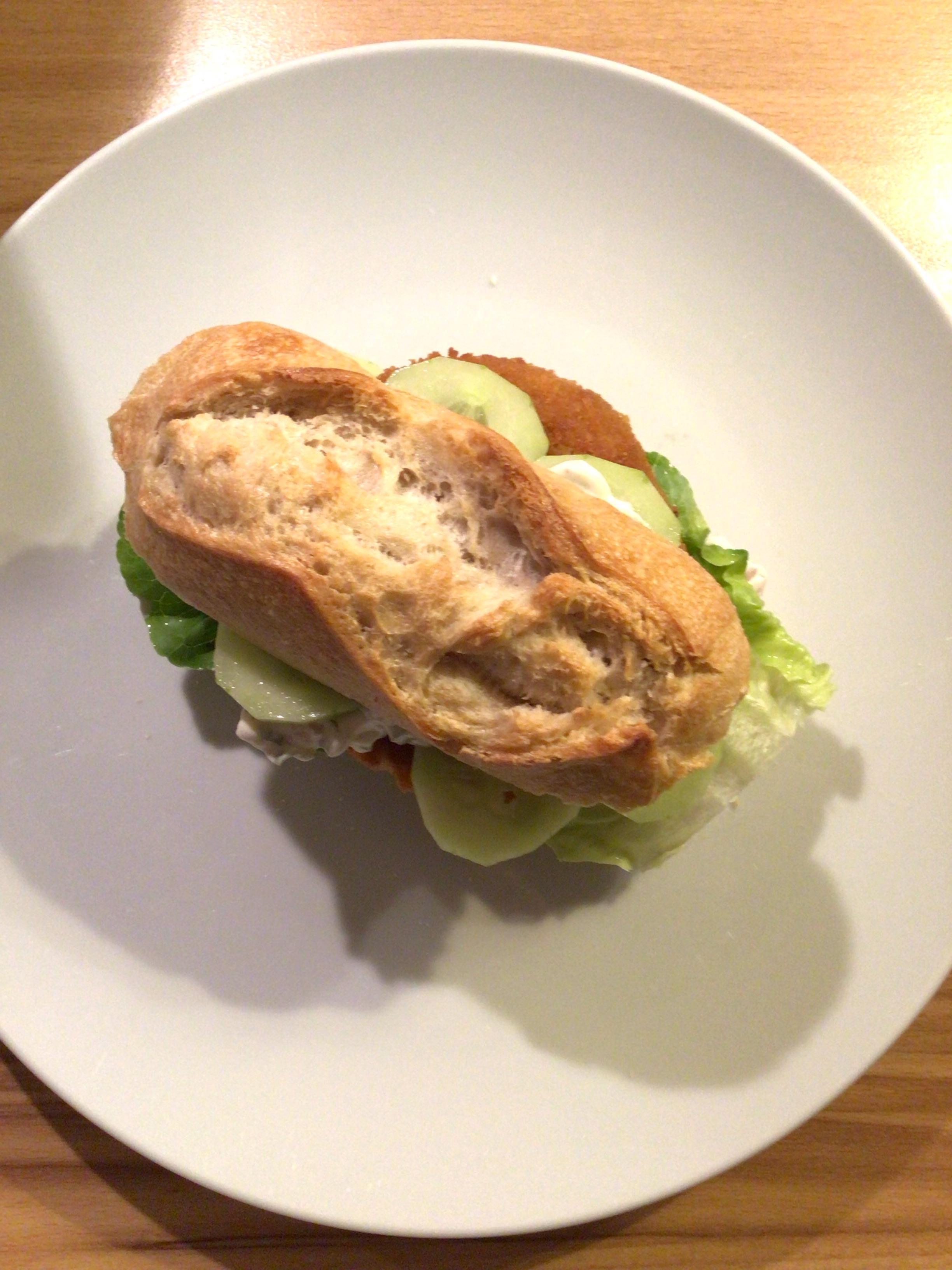 Ein helles, längliches Krustenbrötchen mit der beschriebenen Füllung auf einem weißen Teller, von oben fotografiert. // A light, elongated crust bun with the described filling on a white plate, photographed from above.