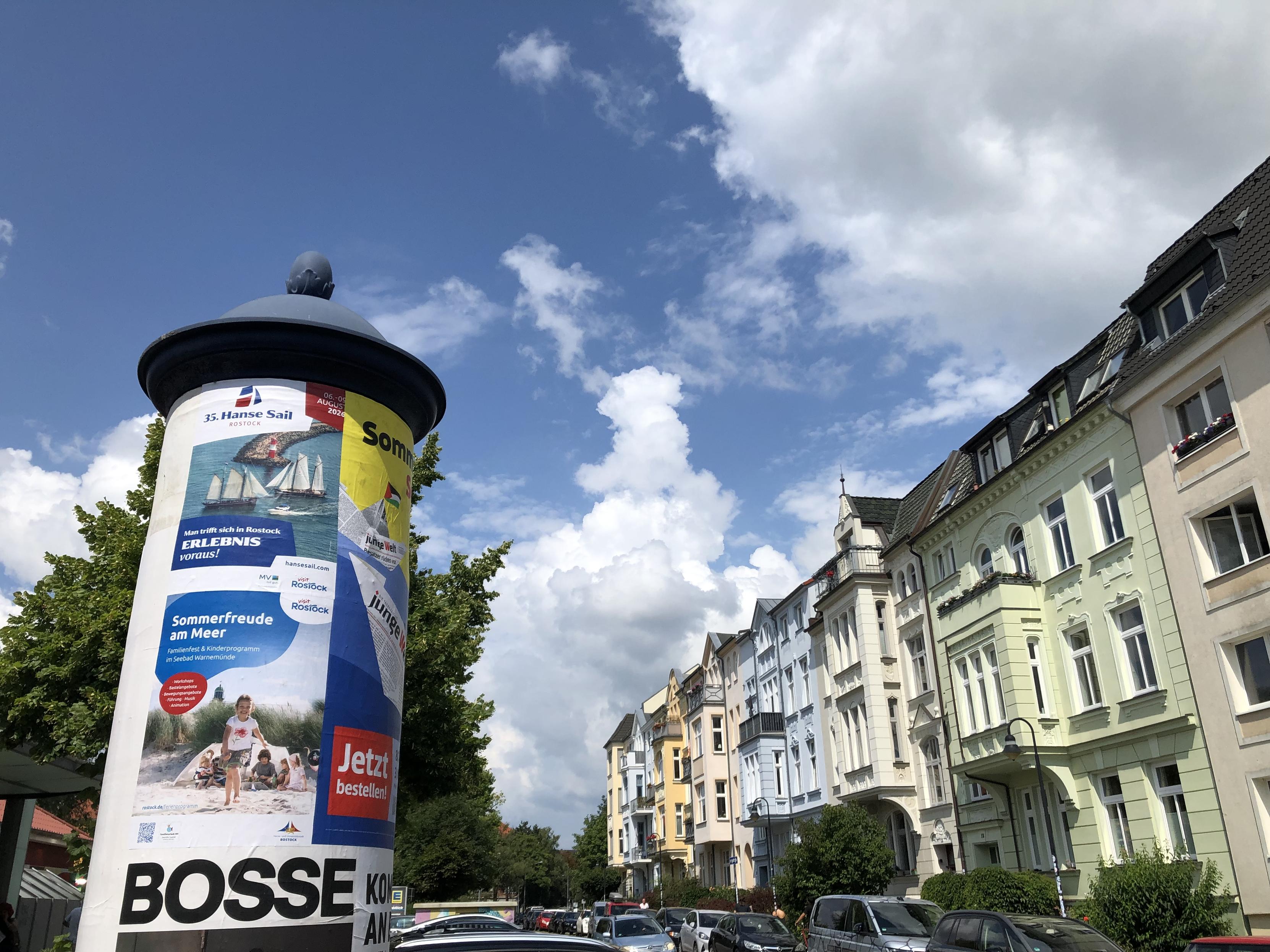 Litfaßsäule vor blauem Himmel mit weißen Wolken und einer Reihe schöner alter verschiedenfarbiger Stadthäuser mit Verzierungen und Erker.