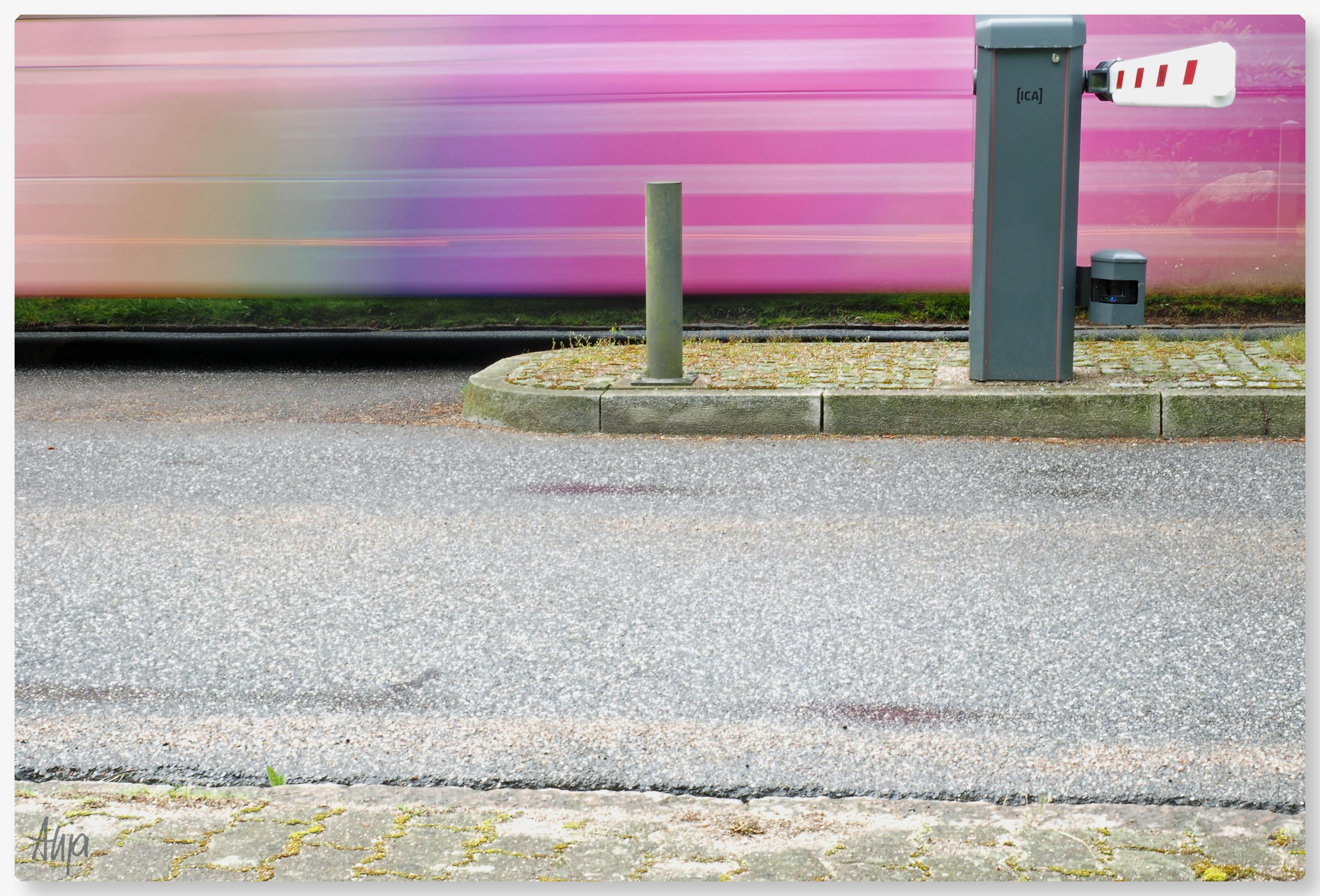 Ein pinkfarbener Bus mit Streifen fährt auf einer Straße an einer Schranke vorbei. Durch eine längere Belichtungszeit ist der Bus verschwommen und nur der Rest des Bildes, graue Straße im Vordergrund und Verkehrsinsel mit Schranke sind im Fokus.

A pink bus with stripes is driving along a road, passing a barrier. Due to a longer exposure time, the bus is blurred and only the rest of the image – the grey road in the foreground and the traffic island with the barrier – is in focus.