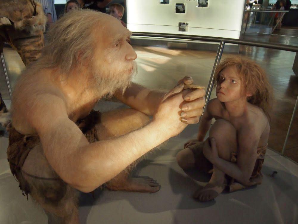 Wachsfiguren. Rekonstruktion von einem Neandertaler Opa, mit einem Neandertaler Kind. Der Alte hockt bei dem jungen. Der junge sitzt daneben. Der Alte hat graues Haar der junge rot blondes. Beide sind sehr hellhäutig, fast Albino. Und beide sind fast nackt.