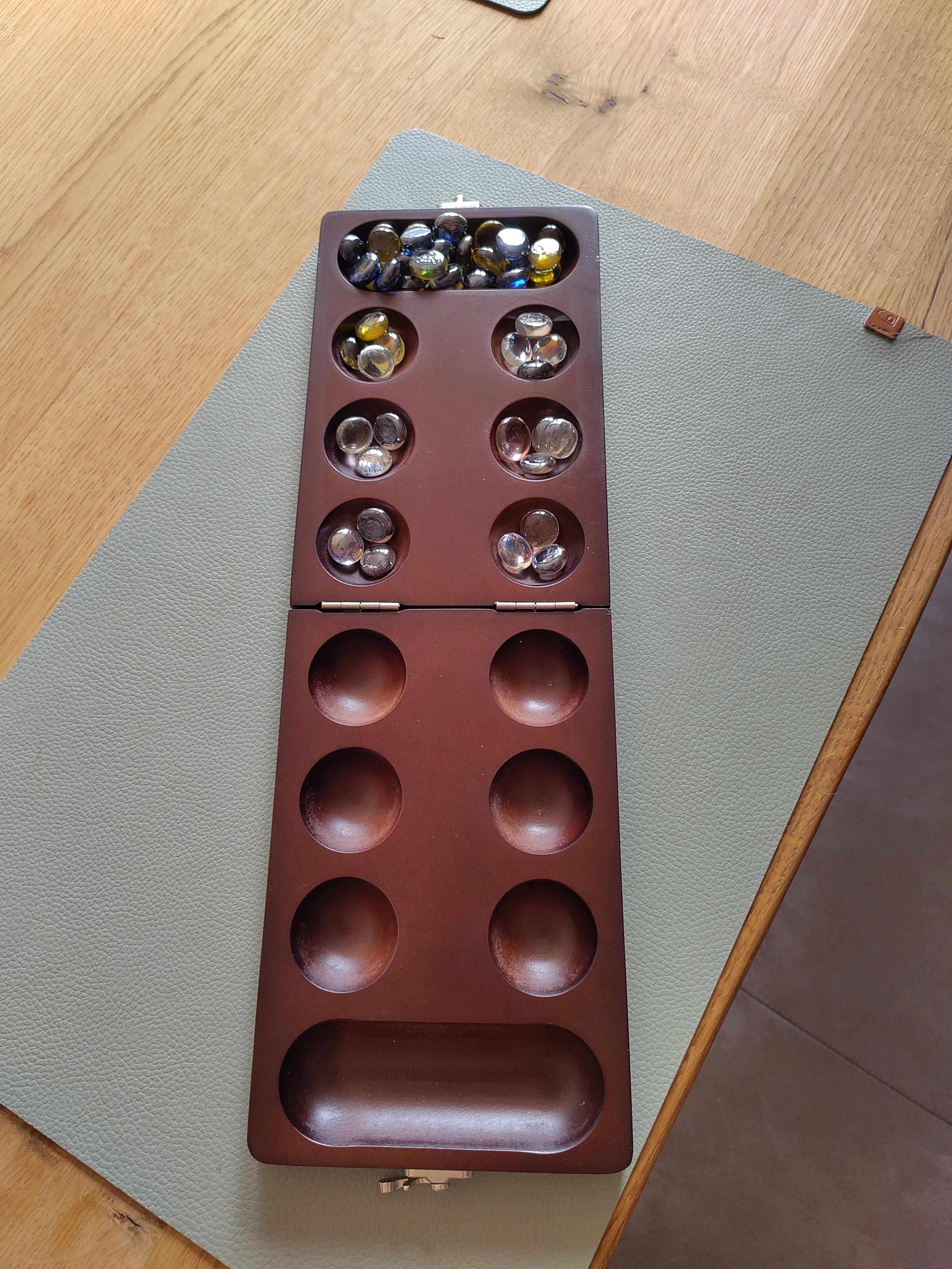 Spielbrett mit 12 kleinen Mulden (je 6 gegenüber) und 2 großen oben und unten. In manchen sind Muggelsteine.