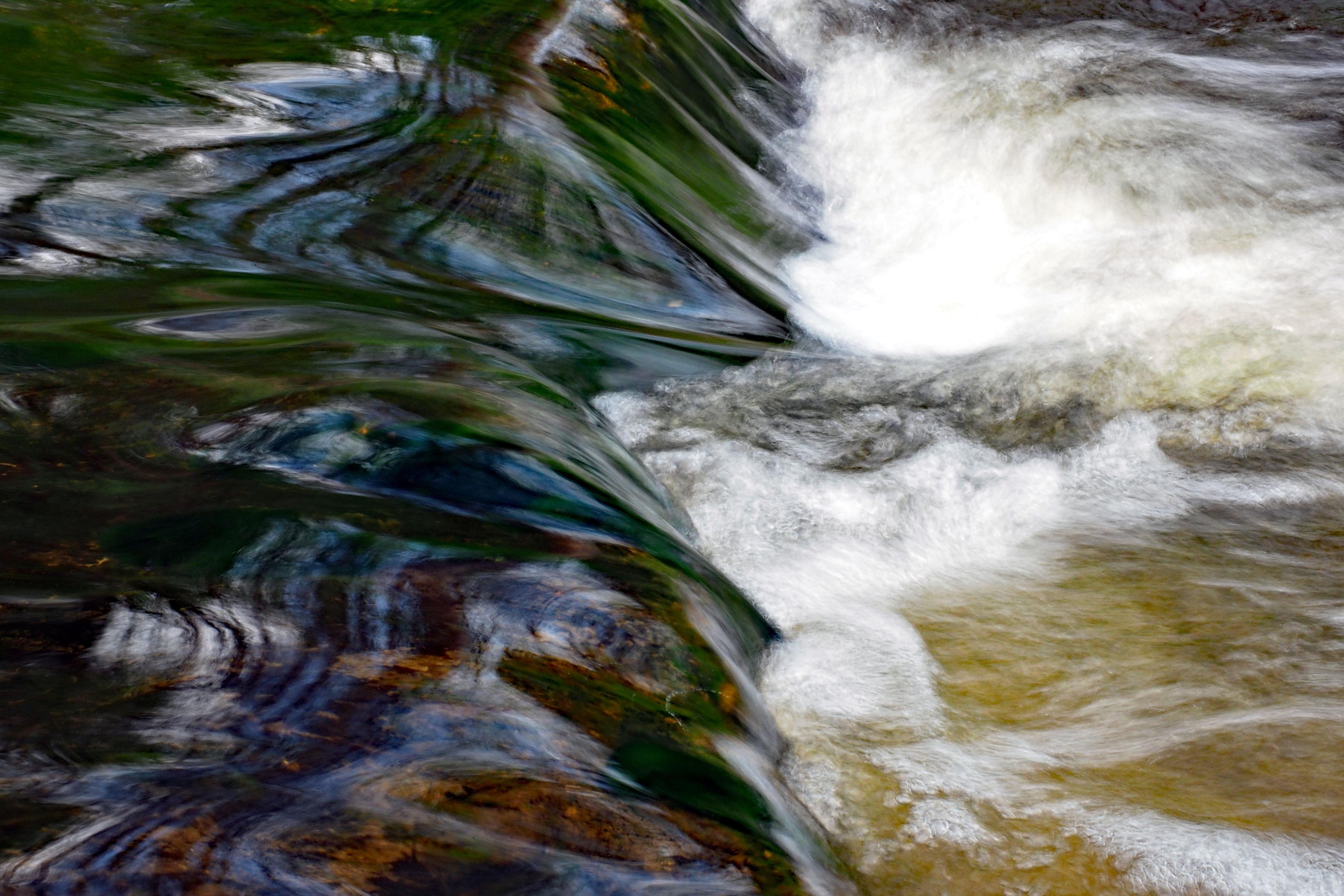 Eine Nahaufnahme eines Flussabschnitts: Das Wasser fließt über eine Felskante. Aufgrund der längeren Belichtungszeit wirkt das Wasser auf der rechten Seite milchig, als wäre es mit einem dicken Pinsel gemalt worden. Auf der linken Seite ist das Wasser dunkel und weist Grün-, Blau- und Rottöne auf, in denen sich das Grün des Ufers spiegelt.

A close-up of a stretch of river: water flows over a stone ledge. Due to the longer exposure time, the water on the right appears milky, as if painted with a thick brush. On the left, the water is dark, with shades of green, blue and red, reflecting the greenery of the bank.