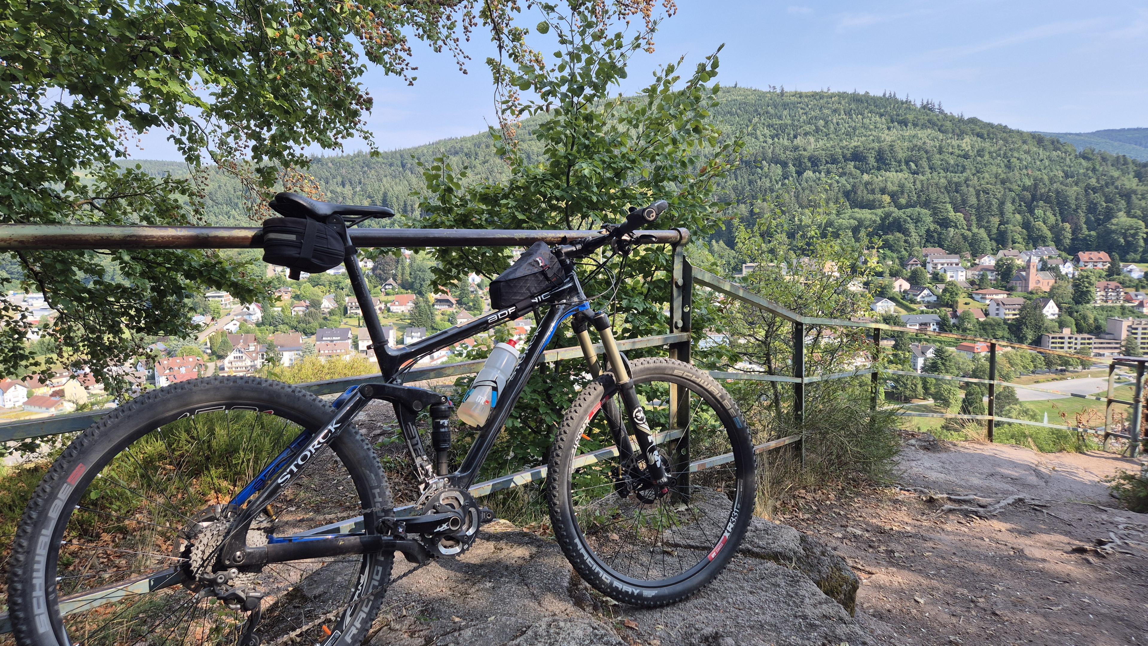 Ein schwarzes Mountainbike lehnt am einem Metall Geländer bei einem Aussichtspunkt der einen weiten Blick über ein malerisches Tal bietet. Im Tal liegt eine Stadt, der Berg auf der gegenüberliegenden Seite ist dicht bewaldet. Über allem ist ein blauer Himmel.