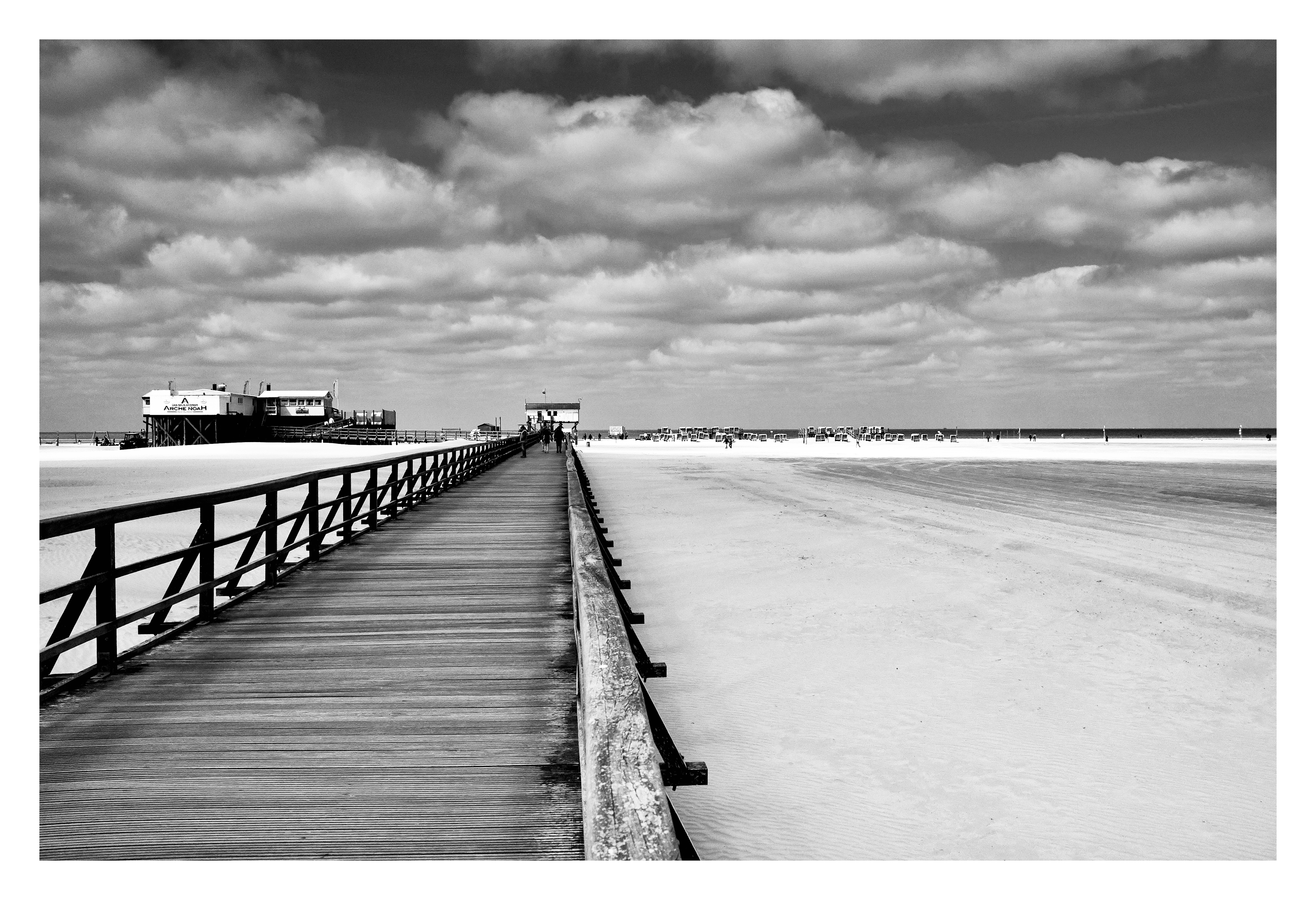 Schwarz-weiß Foto: Im Vordergrund führt die lange Seebrücke über den breiten Sandstrand von St. Peter-Ording in Richtung Meer. Am Ende der Brücke stehen die typischen Pfahlbauten, die auf hohen Stelzen über dem Strand errichtet sind. Dahinter erstrecken sich der weite Strand und das Meer unter einem Himmel mit vielen großen Wolken.