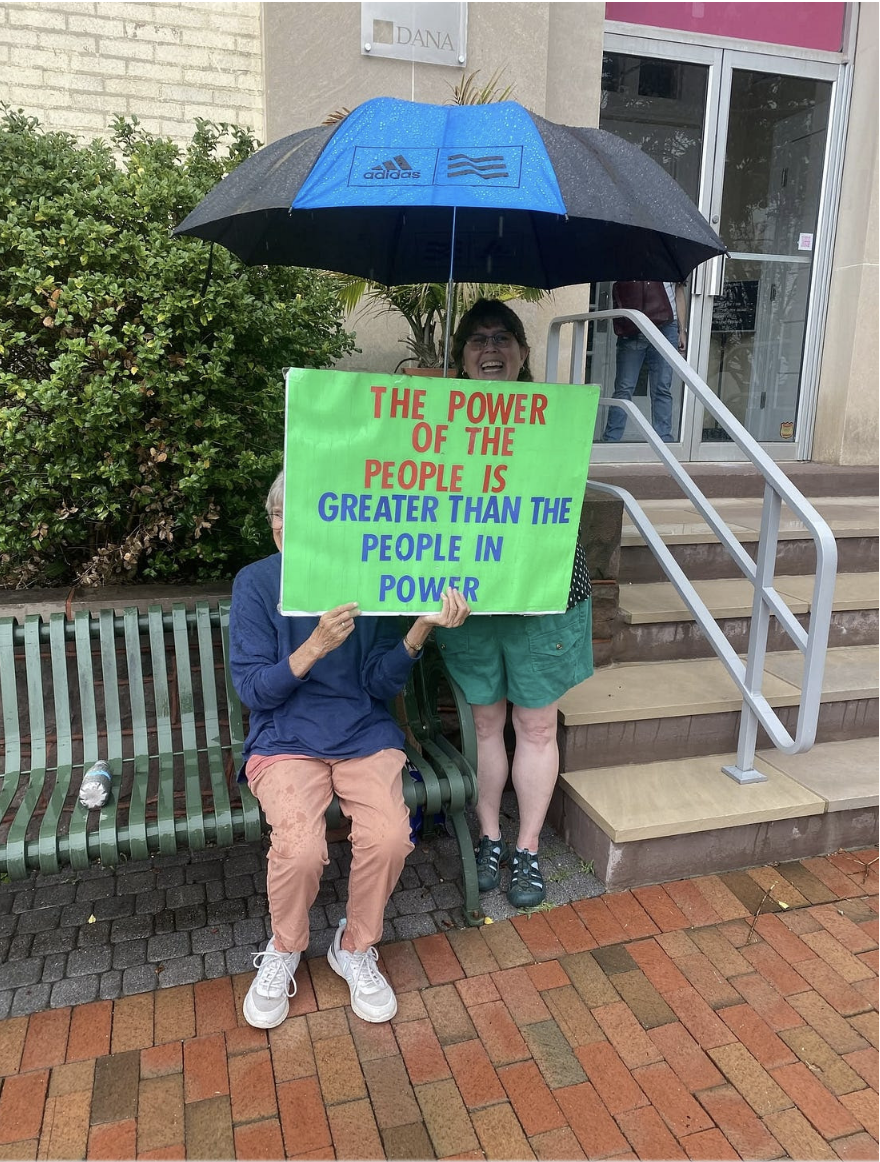 Two protesters in the rain with an umbrella and a sign that says, "THE POWER OF THE PEOPLE IS GREATER THAN THE PEOPLE IN POWER"
