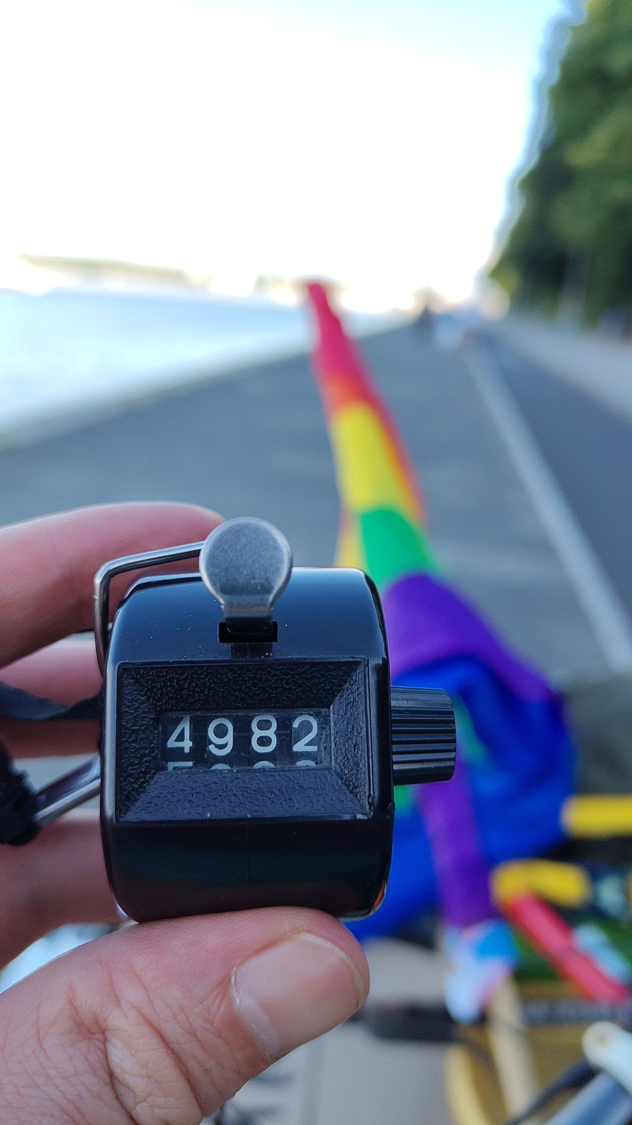 Photo eines Handklickzählers mit der Zahl 4982. Unscharfer Hintergrund mit verschwommener Pride Fahne 🏳️‍🌈