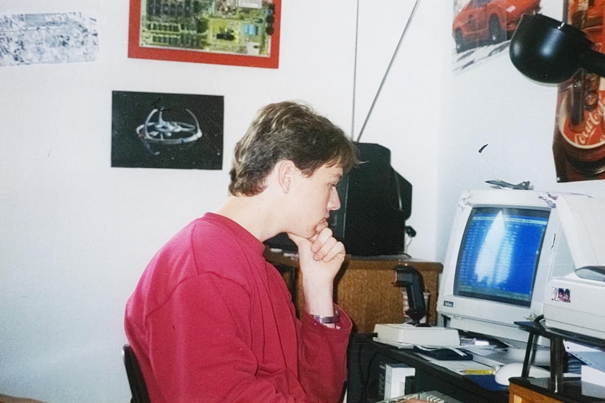 Me, a bit more than 30 years ago, sitting at my desk looking down into the monitor of my first PC. The monitor shows something blue, perhaps Norton Commander or the QBasic editor. On the wall a Star Trek poster, a picture of a Lamborghini and the mainboard of C64 mounted on a red wooden board. On the desk, besides the monitor, a Flight stick from CH and an early Microsoft mouse. In the right partially visible is a dot-matrix printer.