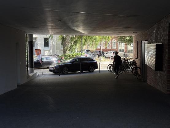 Ein Blick aus einem dunklen, überdachten Bereich auf eine sonnige Straße mit Fahrzeugen und Bäumen. Auf der rechten Seite befindet sich eine Ziegelwand, an der mehrere Fahrräder lehnen und eine Person steht. Im mittleren Bereich ist ein Auto in fast voller Breite im Durchgang zur Straße geparkt.