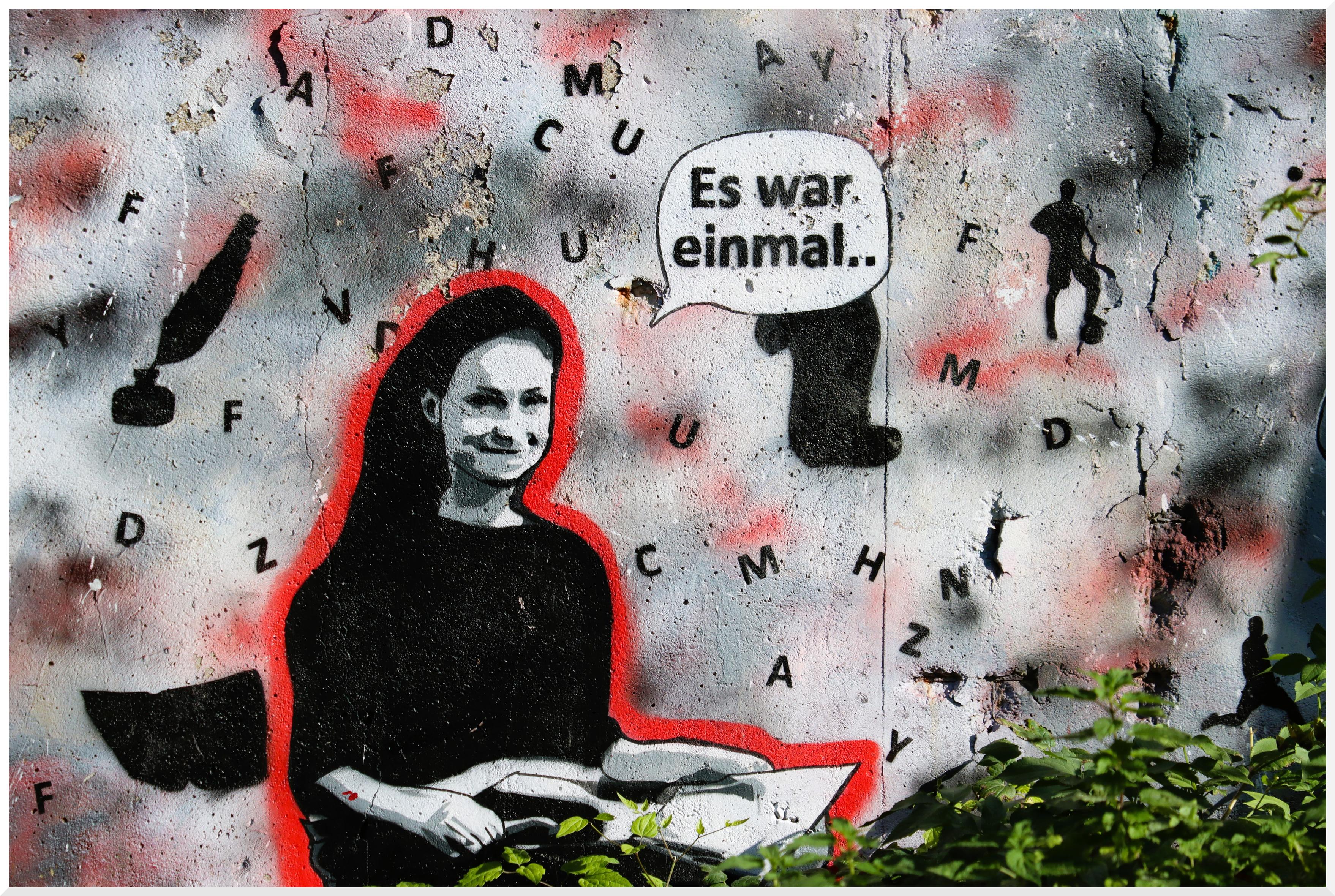 Graffiti an einer Wand. Eine junge Frau mit einem Buch in den Händen. Eine Sprechblase "Es war einmal..". Zahlreiche Buchstaben und einige Objekte wie z.B. eine Feder in einem Tintenfass oder eine Papierrolle.