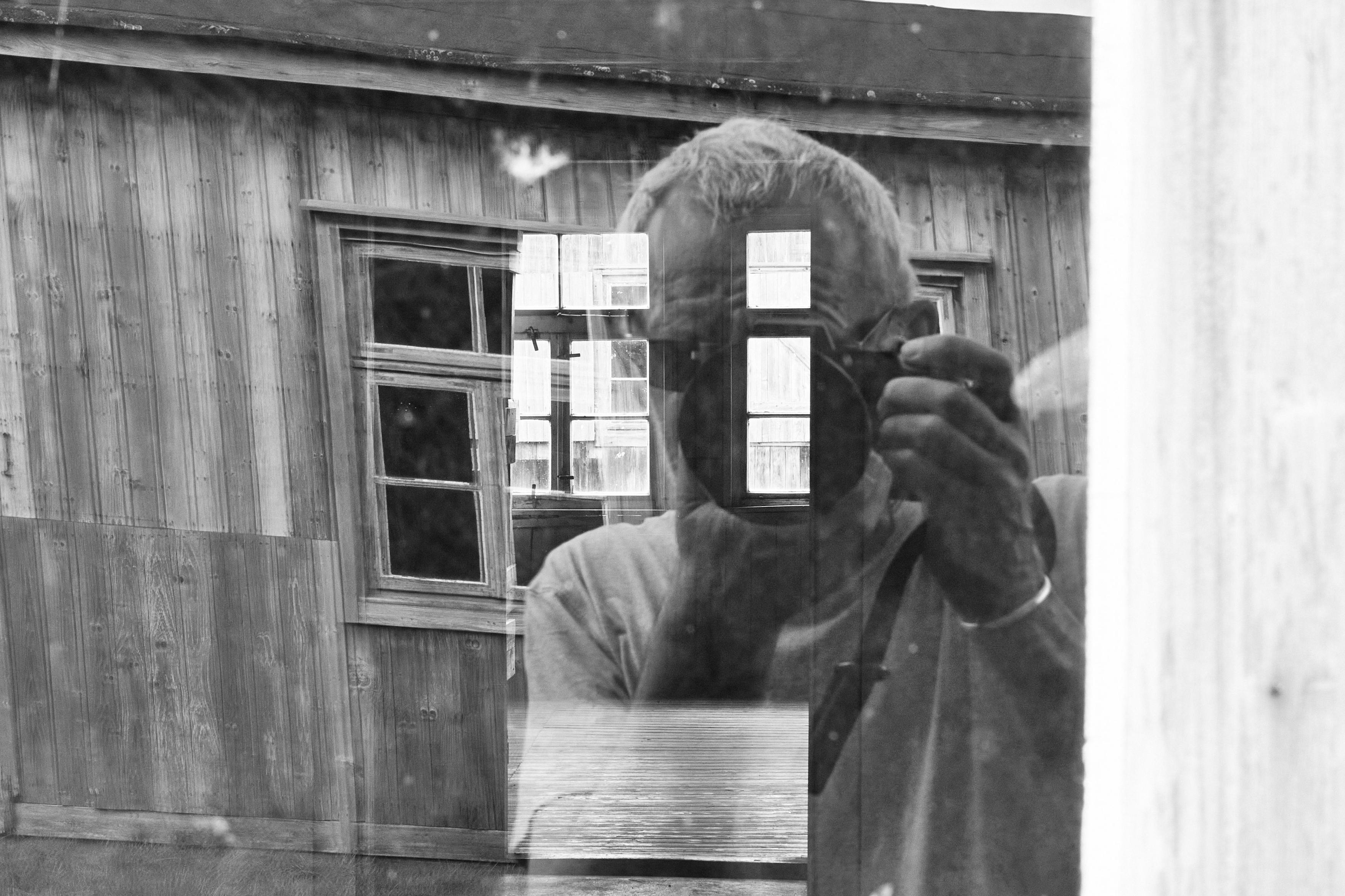 Ein Mann fotografiert durch das Fenster in eine leerstehende Baracke rein. Er spiegelt sich dabei im Fenster. In der Spiegelung ist ebenso eine weitere Baracke in seinem Rücken zu sehen.