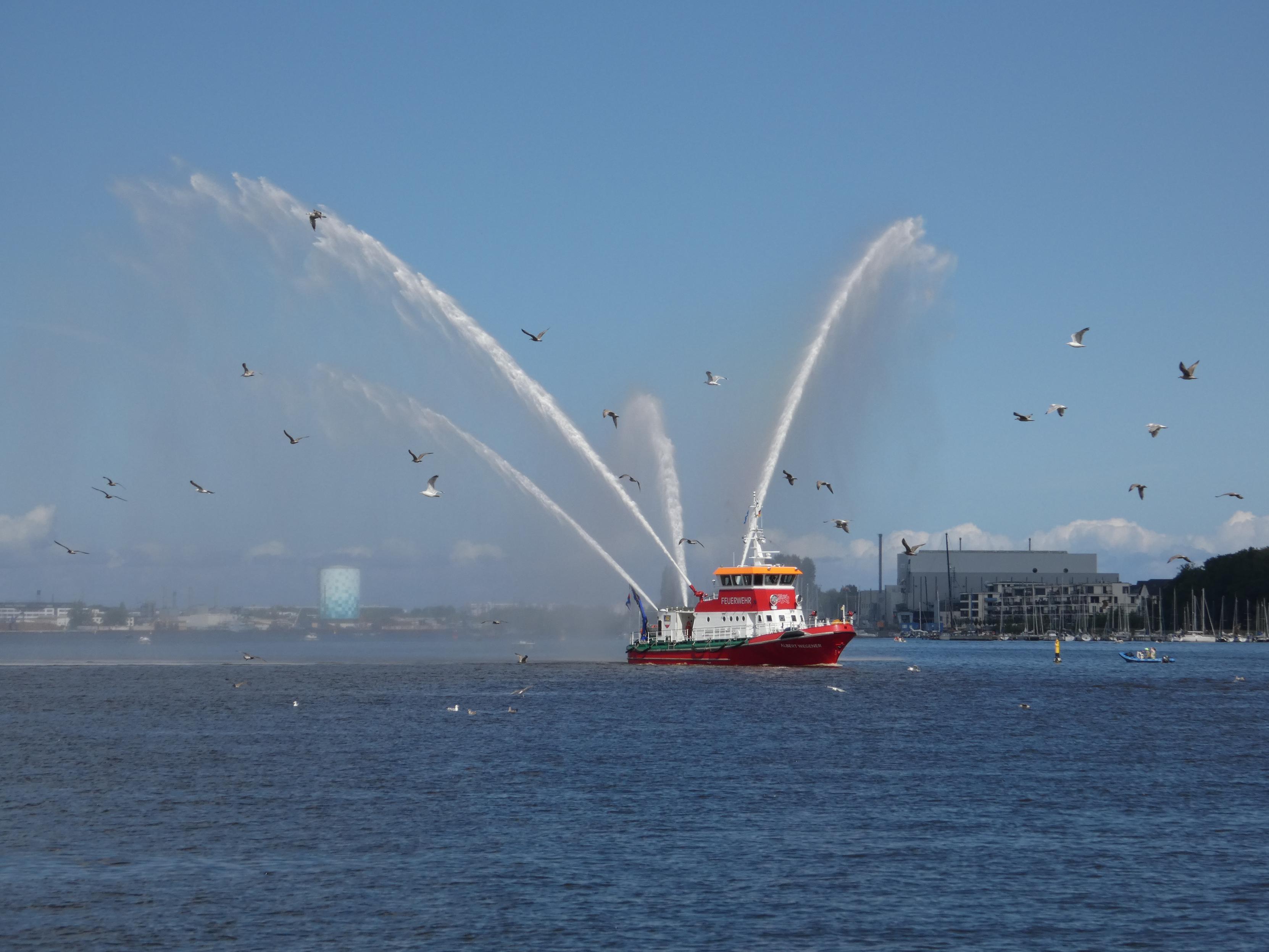 Ein Feuerwehrschiff demonstriert seine Löschkanonen. Vier große, hohe Wasserfontänen werden von dem roten Boot in die Luft gespritzt. Das Feuerwehrboot wird von Möwen umflogen. Im Hintergrund die Warnow und Rostock, Blick nach West/Nordwest.