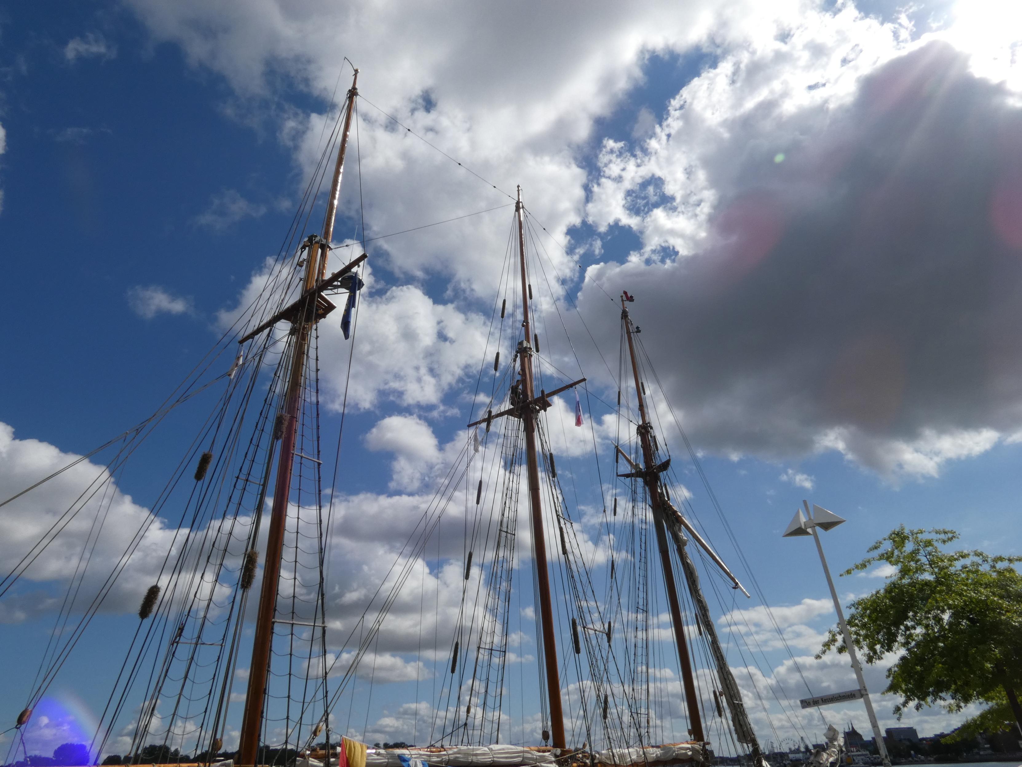 Blick nach oben in den Himmel. Ein dreimastiges Segelschiff. Man sieht die Masten und die Takelage. Wolken, Sonne. Am Warnowufer. Unten ist auch ein Baum zu sehen.