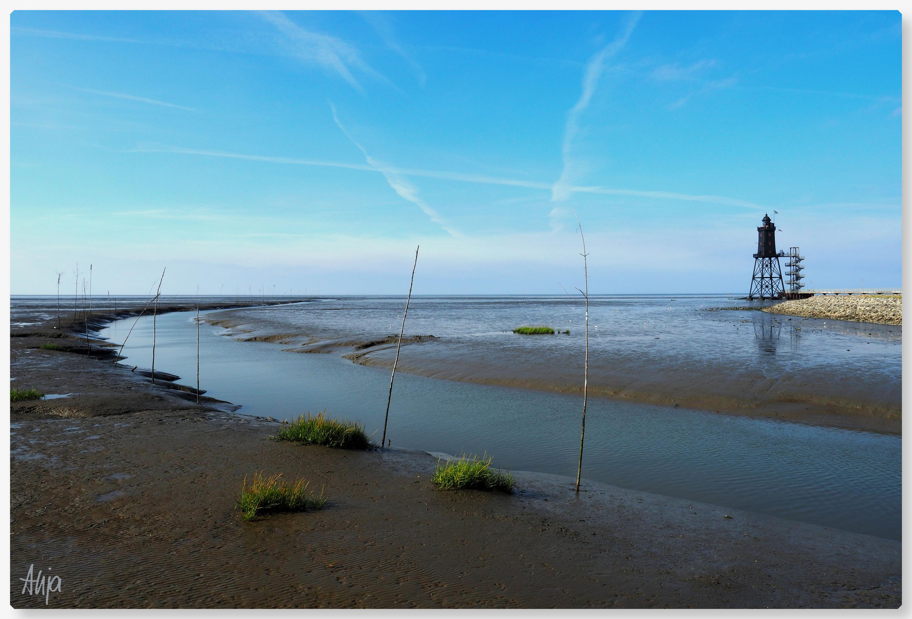 Das Leuchtturmdenkmal bei Obereversand. Im Vordergrund Wasser in einem Priel, abgesteckt mit Birkenzweigen, so genannten Pricken. Der Himmel ist blau, leichte Schleierwolken, das Watt ist braungrau.

The lighthouse monument near Obereversand. In the foreground, water in a tidal channel, marked out with birch twigs, known as ‘Pricken’. The sky is blue with a few wispy clouds; the mudflats are brownish-grey.