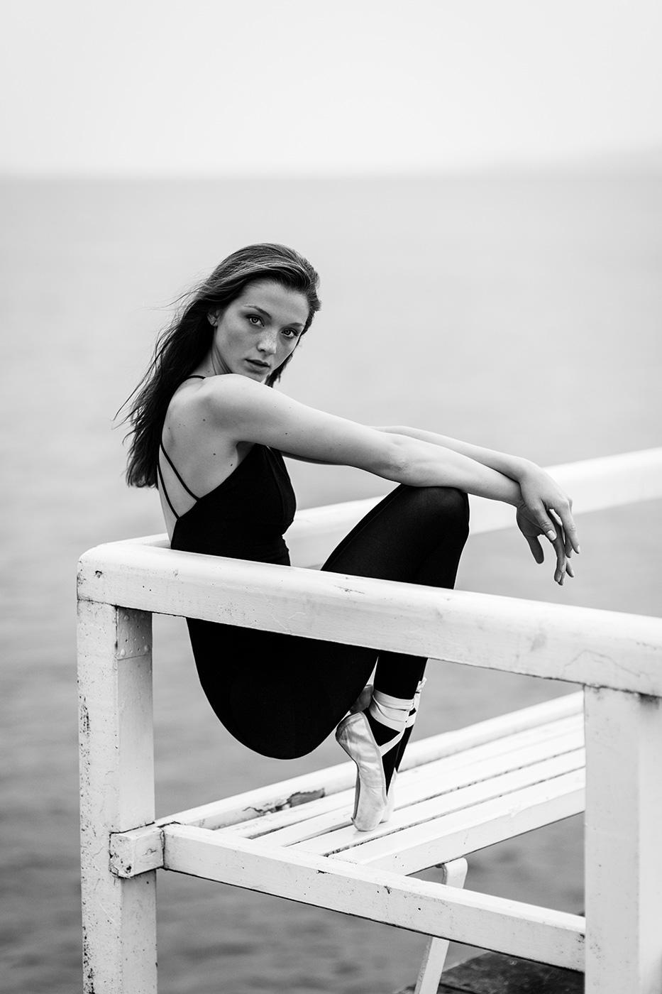 Eine Ballerina sitzt auf einer weißen Bank mit Blick auf den Betrachter. Sie trägt einen schwarzen Tanzanzug und Spitzenschuhe. Ihr Körper ist leicht zur Seite gedreht, die Arme liegen locker auf dem Geländer, die Beine sind ausgestreckt. Der Hintergrund zeigt das ruhige Wasser der Förde, die Szene wirkt ruhig und konzentriert.
