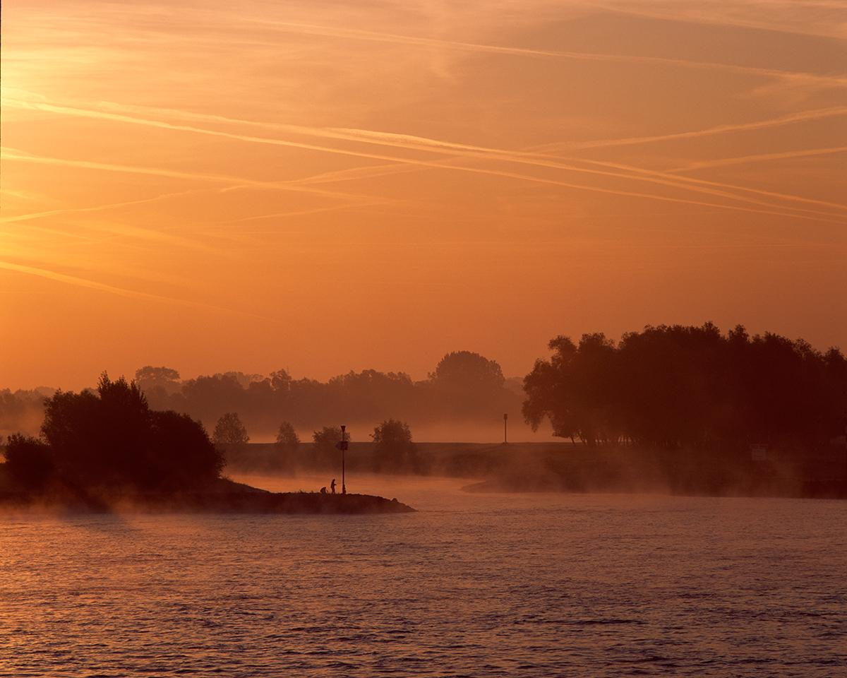 Breiter Fluss mit ruhiger Wasseroberfläche im Vordergrund, im sanften Licht eines orangefarbenen Sonnenaufgangs oder Sonnenuntergangs. Flache Ufer mit vereinzelten Bäumen und niedriger Vegetation, im Hintergrund leichte Nebelschwaden. Am Horizont verlaufen mehrere Kondensstreifen parallel zum Himmel. Die Szene vermittelt die typische Landschaftsstimmung des Niederrheins.