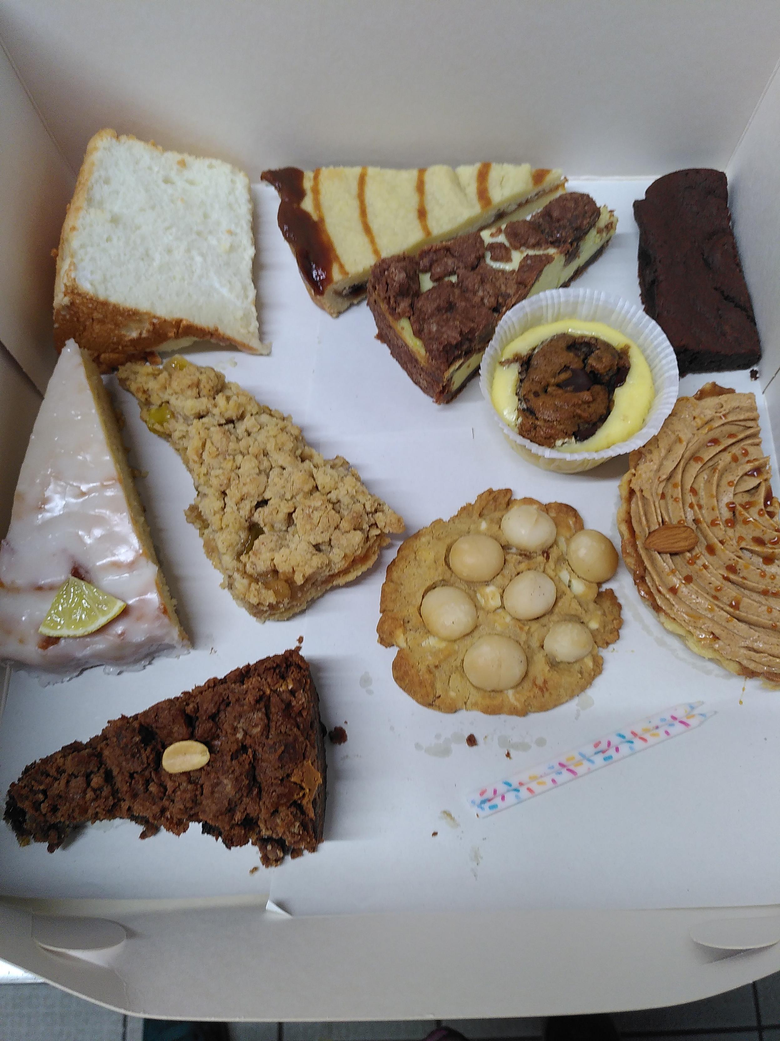 Blick auf zehn verschiedene Kuchenstücke, die in einer weißen Kartonage liegen. Es handelt sich hierbei um folgende Leckereien: oben links ein Stück fluffig-leichter Angel Food Cake, rechts daneben ein Stück Karamellshortbread, ein Stück Zupfkuchen mit Kakaostreuseln (vegan), ein Stück klassischer Schokoladenbrownie; in der Zeile darunter ein Stück Zitronenkuchen (vegan), rechts daneben ein Stück Streuseltarte mit gelben Pflaumen aus meinem Garten (vegan), ein Cheesecake mit eingebackenem Schokokeksteig in der Mitte; in der letzten Zeile ein Stück Bananen-Schoko-Kuchen mit gesalzener Erdnussbutter, ein Handteller großer Keks mit weißer Schokolade und sieben Macadamianüssen und zu guter letzt ein halbmondförmiges Stück Mandelcrèmetörtchen mit Kokoskaramell. Rechts in der Ecke liegen zwei kleine Geburtstagskerzen.
