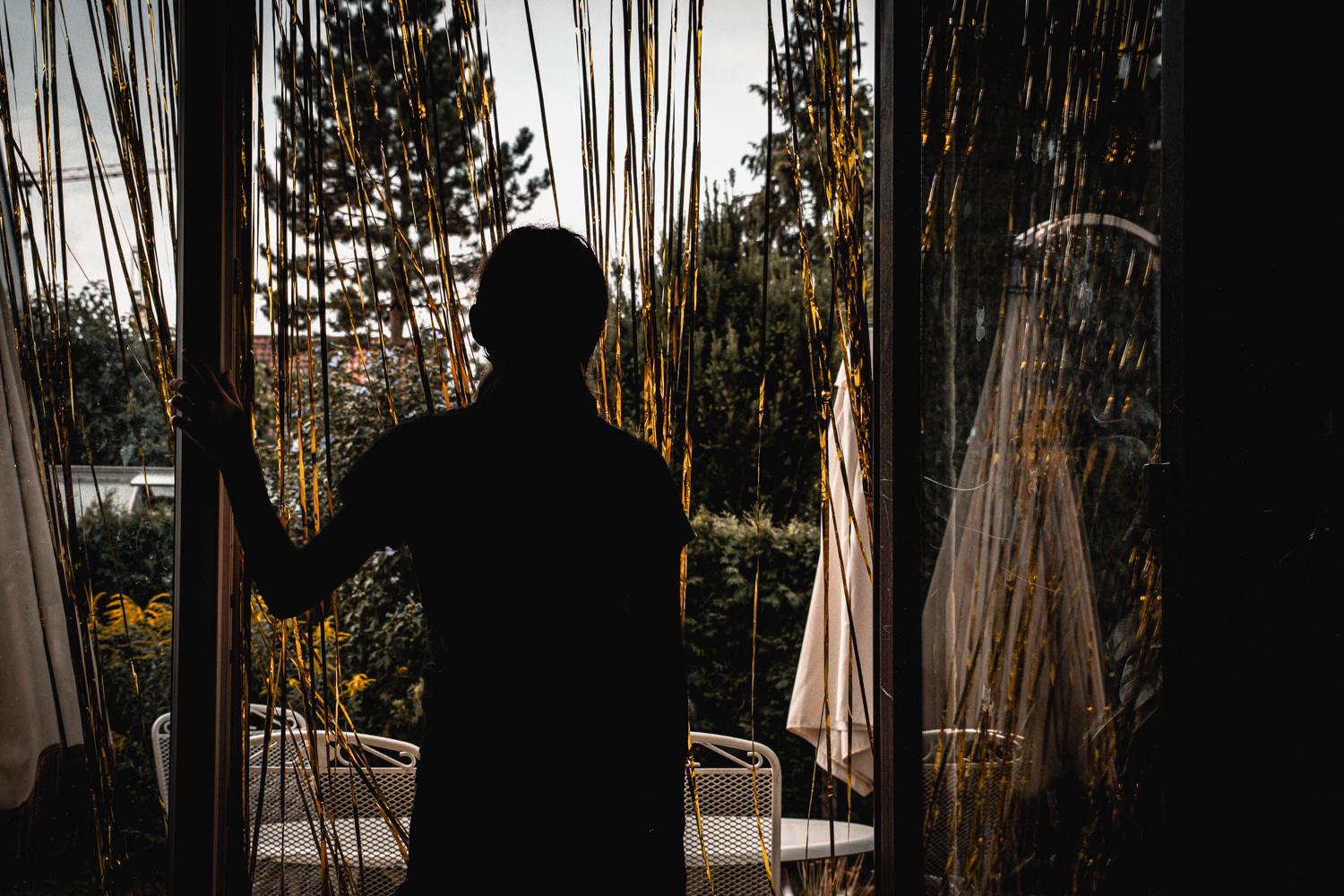 Eine Person steht als Silhouette in einer geöffneten Terrassentür und blickt in einen von Bäumen und Sträuchern umgebenen Garten. Schmale, glänzende Lamellen eines Türvorhangs verlaufen über das gesamte Bild und erzeugen zusammen mit den Spiegelungen auf der rechten Seite eine vielschichtige Bildwirkung. Gartenmöbel und ein geschlossener Sonnenschirm sind im Hintergrund zu erkennen. Die dunkle Darstellung des Innenraums steht in starkem Kontrast zum helleren Aussenbereich und verleiht der Aufnahme eine ruhige, nachdenkliche Atmosphäre.
