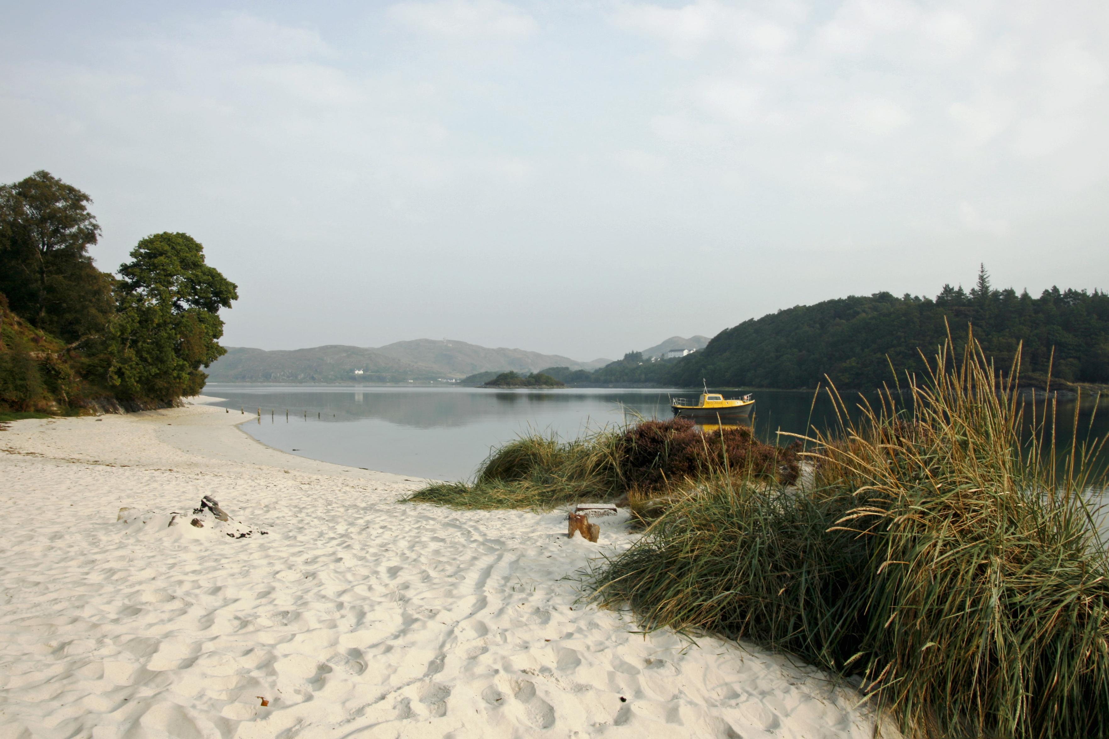 Ein hübscher kleiner weißer Sandstrand mit etwas Dünengras im Vordergrund, Bäumen und Sträuchern links, einem Boot auf dem Wasser sowie Bergen und Hügeln im Hintergrund. Der Himmel ist leicht bewölkt und graublau.

A lovely little white sandy beach with some dune grass in the foreground, trees and bushes to the left, a boat on the water, and mountains and hills in the background. The sky is slightly overcast, grey-blue.