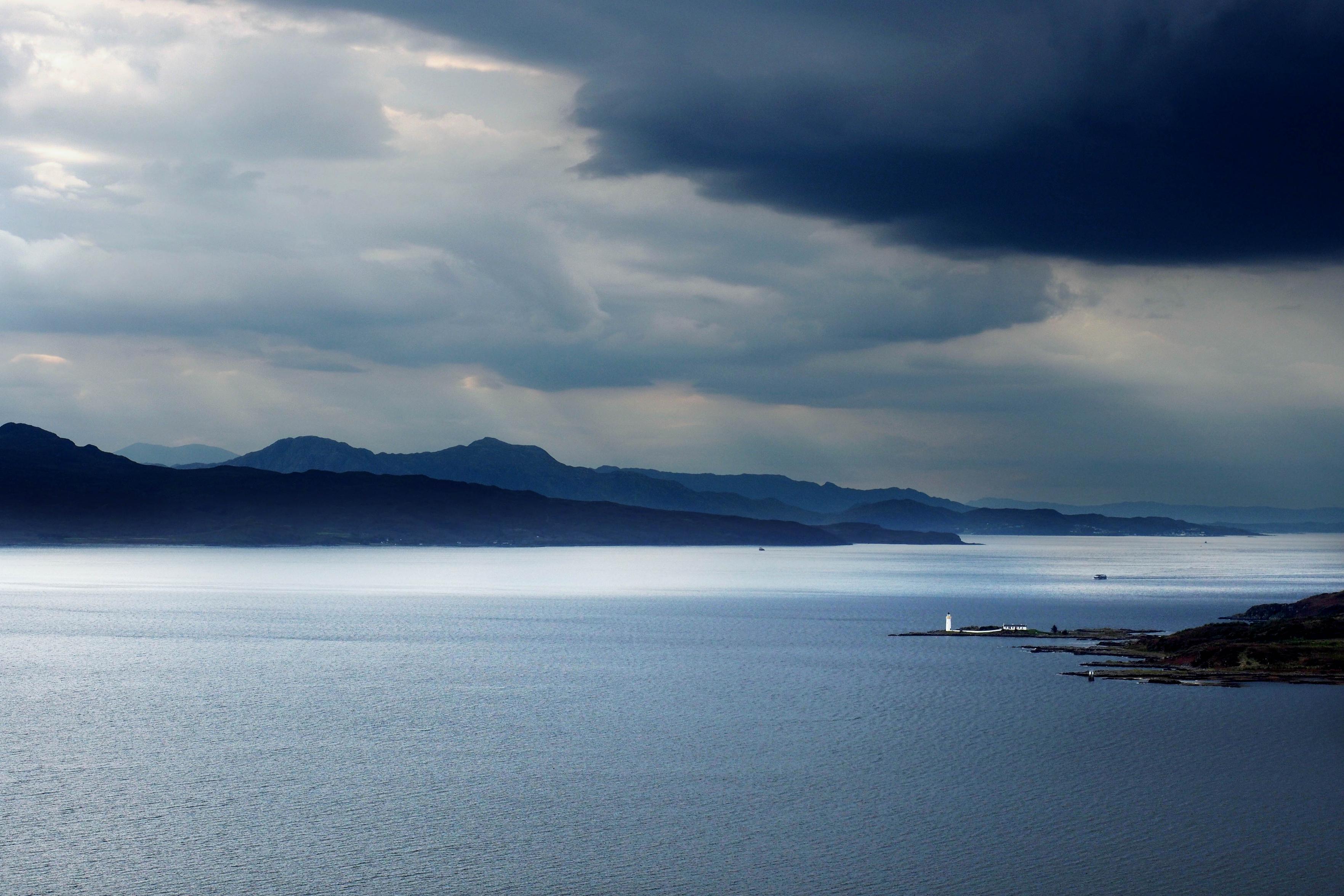 Ein dramatisch wirkender Himmel in Blau, dicke Wolken rechts, über einer Hügelkette und einem Meeresarm. Von rechts ragt eine Landzunge ins Wasser, darauf steht ein weißer Leuchtturm mit weißen Nebengebäuden und einer weißen Mauer, die vom Restlicht der Sonne beschienen werden. Der winzig wirkende Leuchtturm verdeutlicht die Dimensionen dieser schottischen Landschaft.

A dramatic blue sky, with thick clouds on the right, stretching over a range of hills and an inlet. A headland juts out into the water from the right, on which stands a white lighthouse with white outbuildings and a white wall, all bathed in the last rays of the sun. The tiny-looking lighthouse serves to highlight the scale of this Scottish landscape.