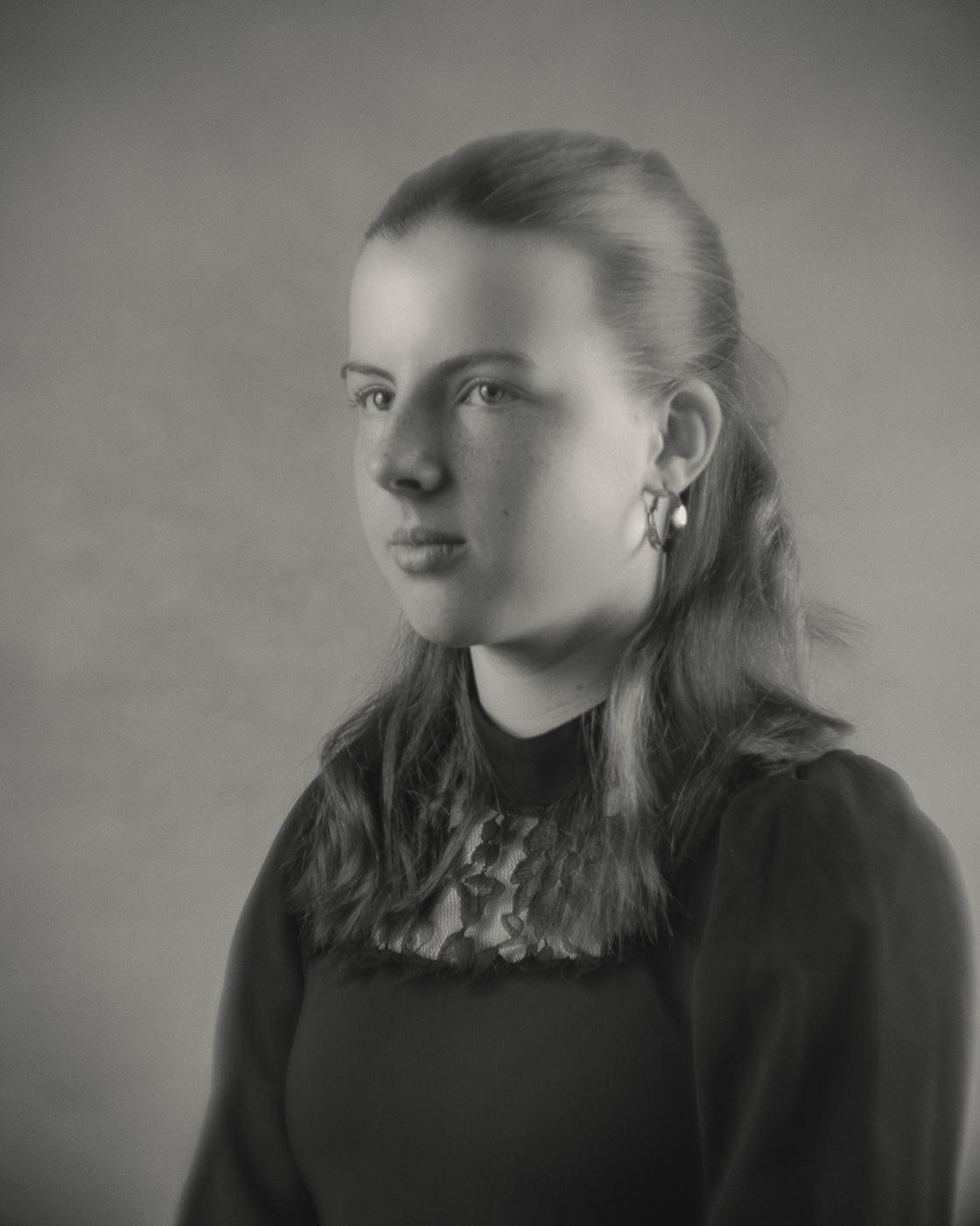 Ein zwölfjähriges Mädchen mit mittellangem, nach hinten gekämmtem Haar blickt leicht zur Seite. Sie trägt ein dunkles Oberteil mit einem auffälligen, gemusterten Kragen und Ohrringe. Das Bild wirkt durch das Weichzeichner-Objektiv sanft und hat einen nostalgischen Charakter.